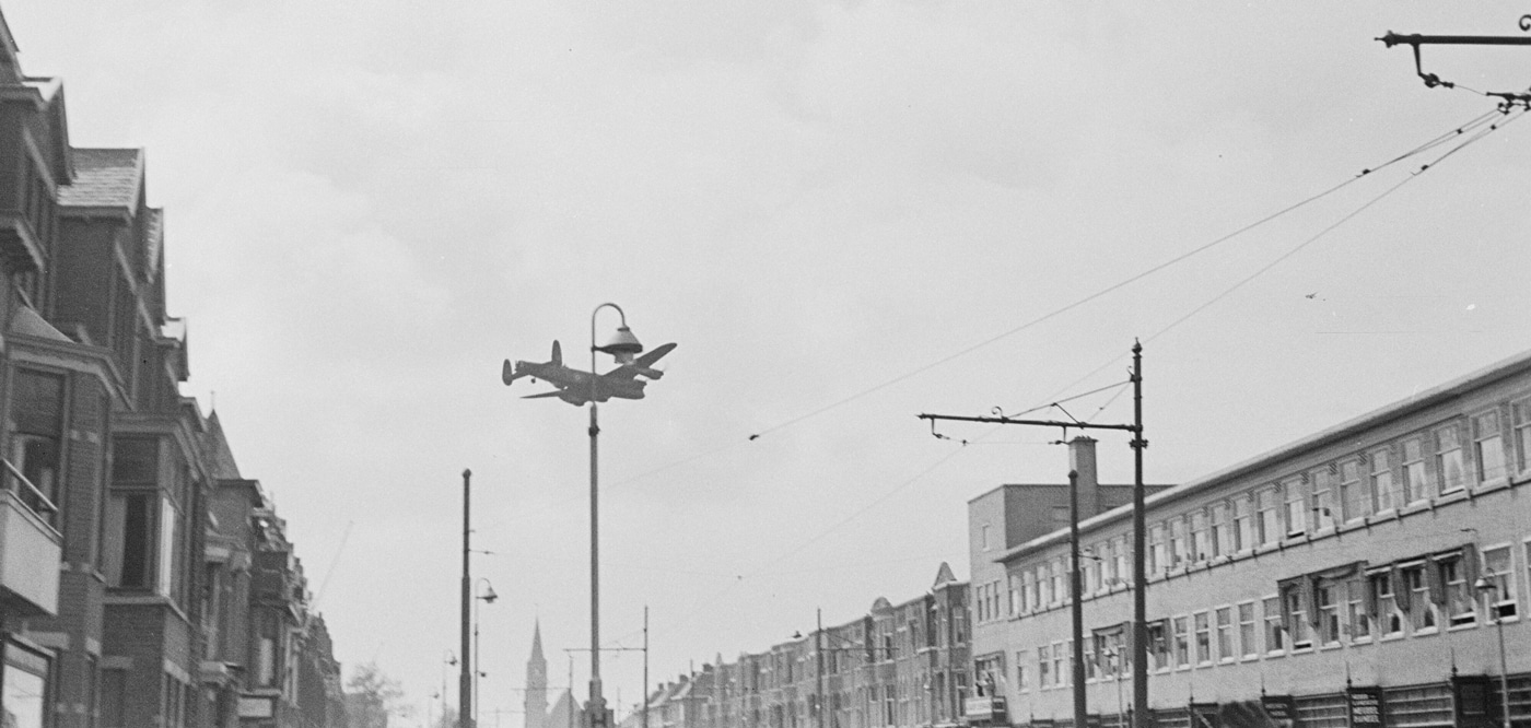 This image shows an RAF Avro Lancaster bomber flying low over Laan van Meerdervoort in The Hague, Netherlands, at the end of World War II as it drops food supplies for Dutch civilians. The aircraft is releasing packages from its bomb bay, which fall toward the city street below. People can be seen on the ground, some looking up or moving toward the dropped supplies, indicating the desperate need for food after years of German occupation. The Lancaster’s four engines and camouflage paint are visible, and the urban setting is marked by rows of houses and a wide street. This mission, known as Operation Manna, was a coordinated effort by the RAF to relieve famine in the Netherlands in spring 1945. The photo powerfully documents the transition from military operations to humanitarian relief at the war’s end. The contrast between the bomber’s original combat role and its use in food delivery is central to the scene.