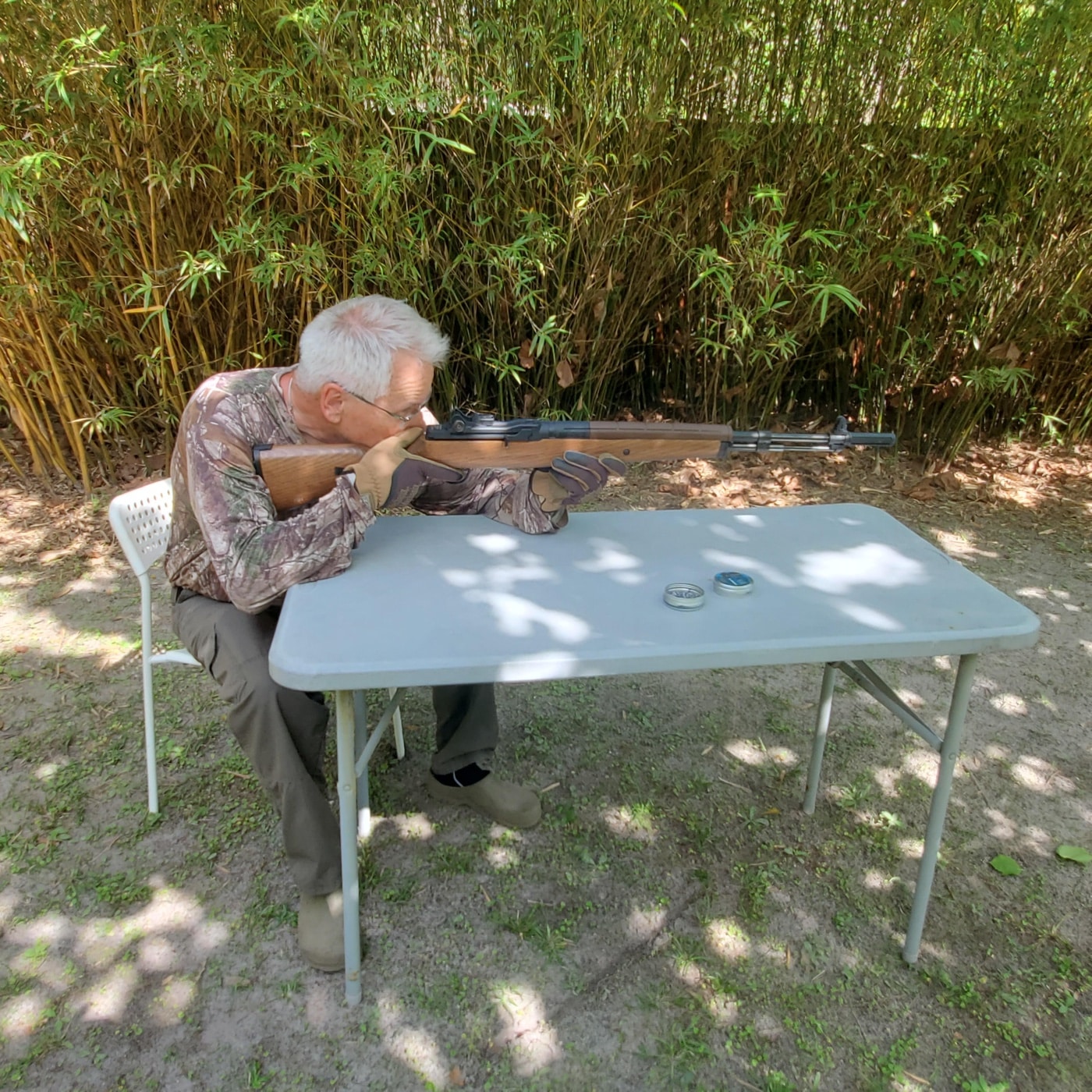 The author is pictured seated at an outdoor table, aiming the Springfield M1A air rifle toward a distant ROJO RJ4 Tactical Target. The table is used as a steady shooting platform, with pellets and shooting accessories laid out nearby. The author is focused on the sights, demonstrating a typical shooting position for target testing. In the background, the ROJO target is visible, set up for accuracy testing as described in the Rojo RJ4 Tactical Target review. This scene gives a realistic look at how the product was field tested with the actual rifle and target setup.
