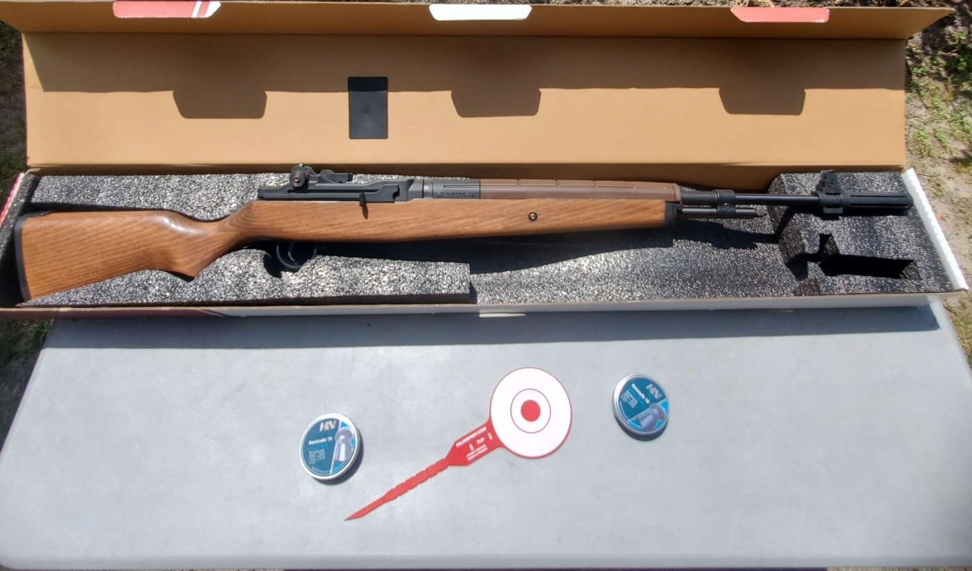 A Springfield Armory M1A air rifle is positioned next to a tin of .22 caliber pellets and a ROJO RJ4 Tactical Target. The rifle’s wooden stock and metal barrel are clearly visible, and the pellets are shown in an open container with several loose rounds nearby. The ROJO target is set out flat or propped up, displaying its red bullseye and target rings. This arrangement gives a straightforward view of all the gear used in the Rojo RJ4 Tactical Target review, making it easy to see what was tested. Useful for readers who want to see the combination of rifle, ammunition, and target used in the field tests.