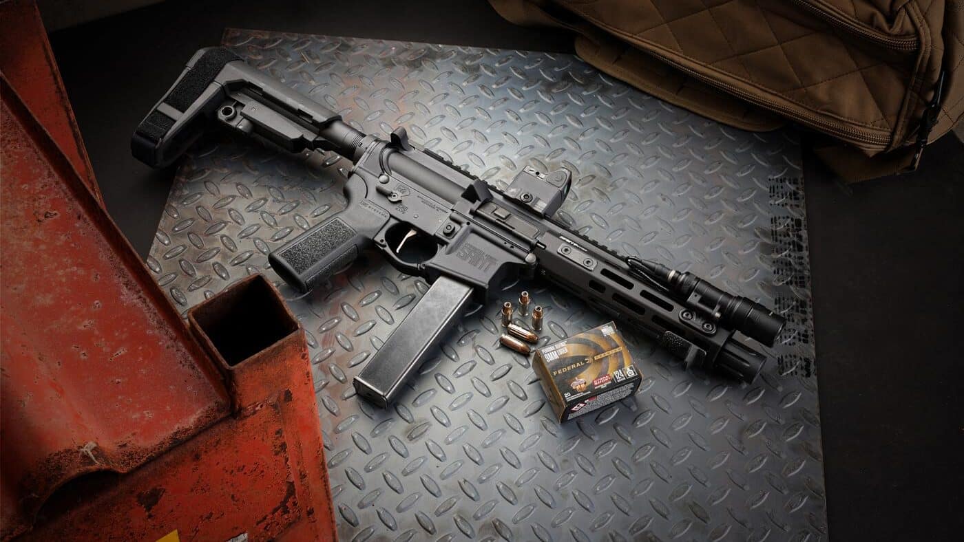 A Springfield Armory SAINT Victor 8.5” pistol chambered in 9mm is displayed next to a range bag and several boxes of Federal defensive ammunition. The pistol features an M-LOK handguard, SB Tactical SBA3 brace, and a distinctive Muzzle Drum. Its overall configuration reflects a setup ideal for home defense or range use. This layout showcases key accessories commonly used with AR-pattern pistols in pistol calibers. The image helps illustrate the practical use-case discussed throughout the SAINT Victor 8.5” 9mm Pistol review.