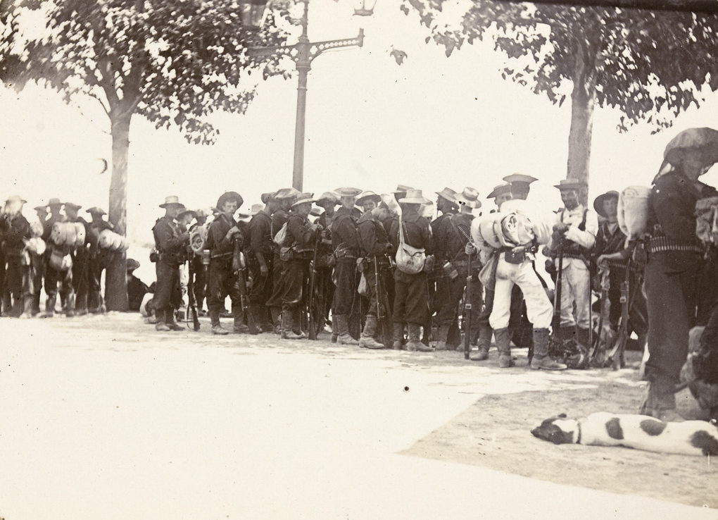 The photograph shows a busy encampment as members of the Seymour Expedition prepare to depart on their mission to relieve the besieged Legation Quarter in Peking during the Boxer Rebellion. Soldiers from different nations — including British, American, Japanese, Russian, and other allied contingents — are visible assembling wagons, organizing supplies, and readying horses or mules for the journey. Officers consult maps or give instructions while enlisted men pack tents, ammunition, and food into transport wagons. The setting is a temporary camp on open ground or the outskirts of a Chinese city, with makeshift shelters, stacks of gear, and flags from multiple nations in view. The image captures the urgency and complexity of organizing a large multinational force for a rapid military advance under uncertain conditions. The presence of so many different uniforms and languages highlights the international scope of the operation. The photo offers insight into the logistical challenges and teamwork required for such an expedition during the Boxer Rebellion.