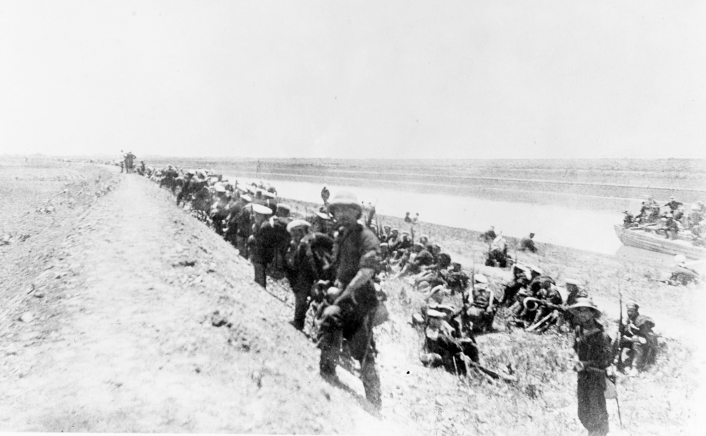 The photograph shows a tense scene along the embankment of the Pei-Ho river during the retreat of the first Peking relief expedition in 1900. In the foreground, British bluejackets (Royal Navy sailors) crouch or lie low behind the earth embankment, some aiming rifles while others tend to the wounded or pass ammunition. To the right, a large Chinese junk is pulled up to the riverbank, loaded with wounded soldiers and crates of medical supplies or food. In the background, an American military detachment is visible, similarly using the embankment for cover. On top of the embankment stand a group of officers, including Admiral Seymour of the Royal Navy and Commander McCalla of the U.S. Navy, overseeing the situation and possibly signaling orders. The atmosphere is tense, with signs of recent shelling — smoke, debris, or soldiers taking cover — indicating that the area is under fire from Chinese artillery. The distant landscape shows open fields where Chinese cavalry are reportedly preparing to attack, though not visible in the image. The photo conveys the urgency, coordination, and hazards faced by the international relief column as it attempted to escape encirclement near Yant Sun and the Siku arsenal during the Boxer Rebellion.