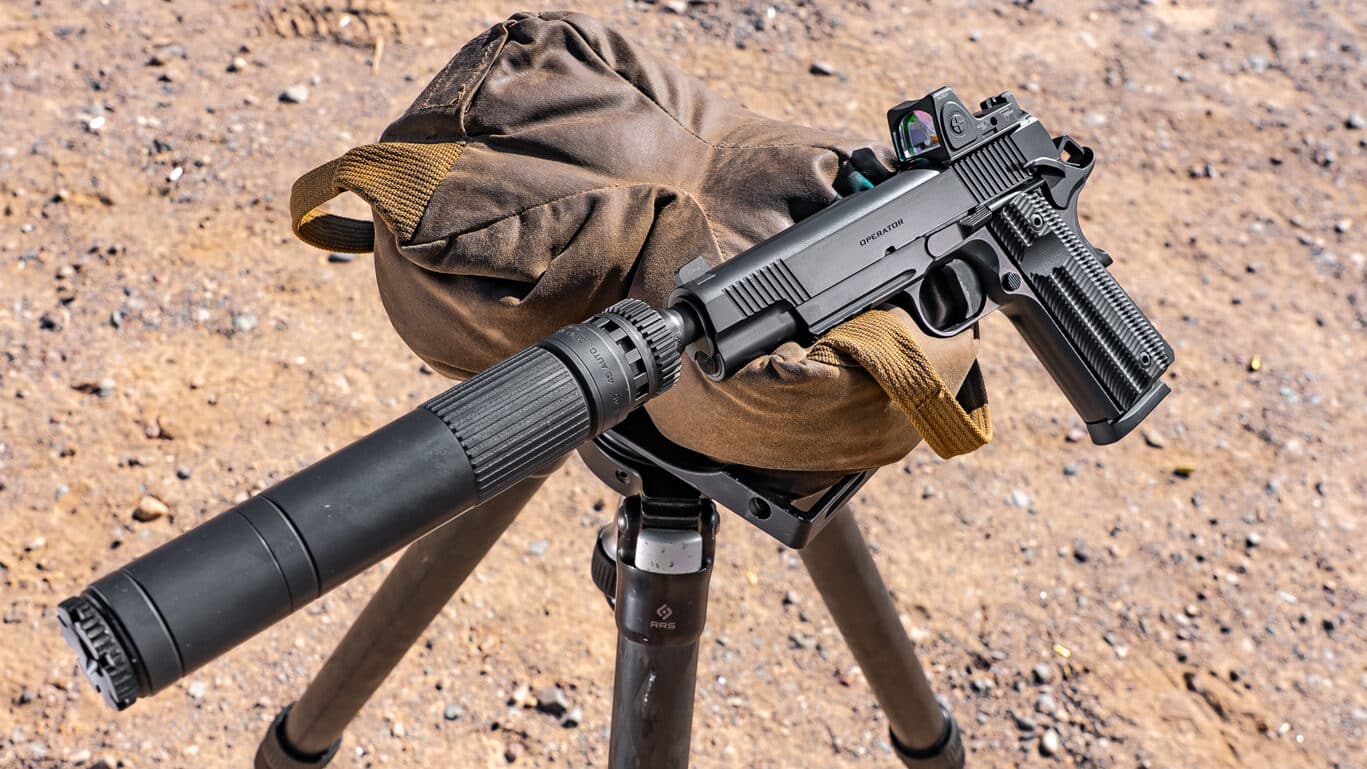 Image shows the Springfield Armory 1911 Operator AOS Threaded .45 ACP displayed on a shooting range surface. The handgun is not being handled; it’s laying flat, fully outfitted with a threaded barrel, a suppressor attached to the muzzle, and a red dot sight mounted on the slide. The Cerakote finish gives the pistol a modern, durable appearance. The overall setup demonstrates compatibility with both sound suppressors and optics, appealing to tactical shooters and enthusiasts. The image composition is clear and professional, making all key features visible, from the accessory rail to the controls and the profile of the suppressor. This photo would be useful in an article discussing upgrades, handling, and performance of the 1911 Operator AOS. Optimized for keywords like “1911 Operator AOS Threaded .45 ACP,” “Springfield Armory,” “suppressor-ready 1911,” and “optics-ready .45 ACP pistol.”