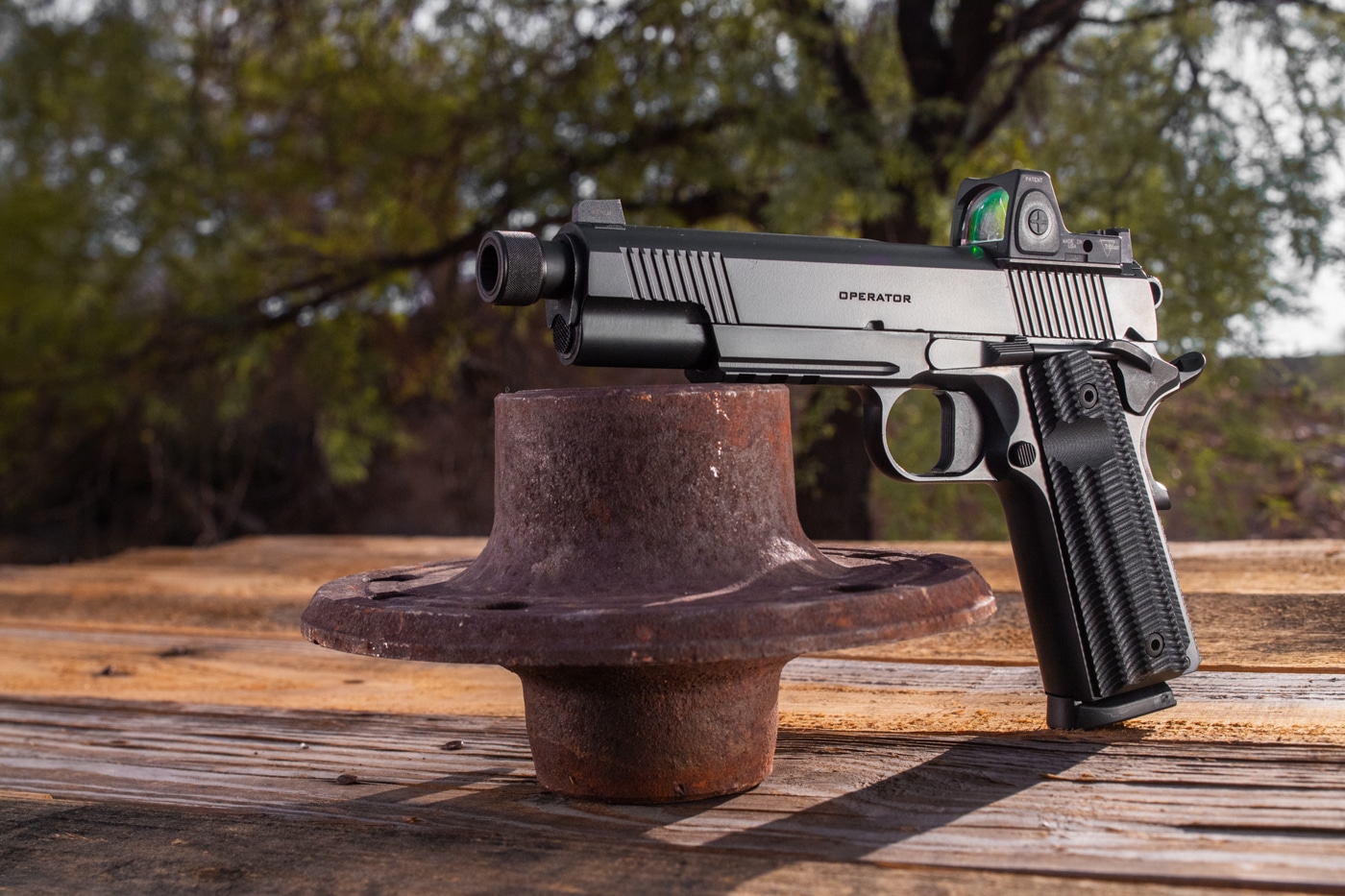 The image presents a Springfield Armory 1911 Operator AOS Threaded .45 ACP lying on an outdoor table, with natural lighting making all details clear. The handgun’s barrel is fitted with a knurled thread protector instead of a suppressor, showing off its threaded design while maintaining a streamlined look. A Trijicon red dot optic is mounted on the slide, emphasizing the pistol’s Agency Optic System (AOS) capability. The Cerakote finish and tactical controls are clearly visible. No hands or accessories are present; the focus is entirely on the pistol and its main features. The outdoor setting provides a casual yet informative context for range or field use. Keywords optimized for this ALT text include “Springfield 1911 Operator AOS,” “thread protector,” “Trijicon red dot sight,” and “optics-ready .45 ACP.”