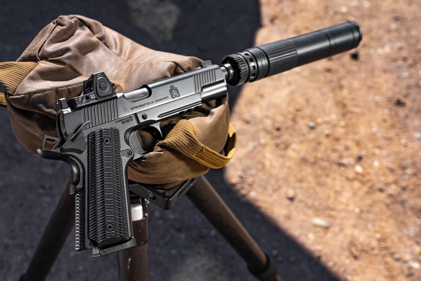 The photo shows the Springfield Armory 1911 Operator AOS Threaded .45 ACP lying on a shooting bench at the range. A red dot sight is mounted on the slide, and a full-size suppressor is attached to the threaded barrel. The pistol is not being held; it’s positioned so the entire length, from muzzle to grip, is easy to see in one shot. Both the optic and suppressor stand out, illustrating how the firearm looks when set up for actual range use. There are no hands, ammunition, or other accessories visible, keeping the attention on the gun itself. The background includes subtle range details — such as target berms or spent brass — but is mostly out of focus, highlighting the pistol as the main subject. This image gives a realistic impression of the gun’s overall footprint and how all the major upgrades work together.