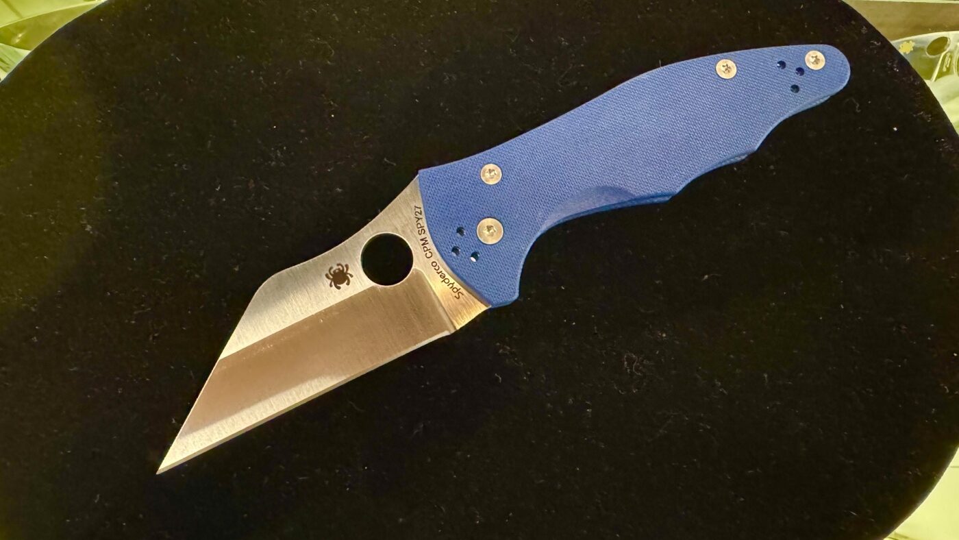 This image shows the Spyderco Yojimbo 2 Cobalt Blue folding knife on display at Blade Show 2025, with its Wharncliffe blade open. The blade is made from CPM SPY27 steel, and its straight cutting edge and fine tip are clearly visible. The handle is crafted from cobalt blue G-10, giving the knife a striking appearance that stands out among other models. The compression lock mechanism is visible near the pivot, and the pocket clip is attached to the handle. The knife is set on a Spyderco-branded display mat, with other Spyderco models and promotional materials nearby. Booth lighting brings out the vivid blue handle and the satin finish on the blade. The photo highlights the design details and materials of this new Yojimbo 2 release.