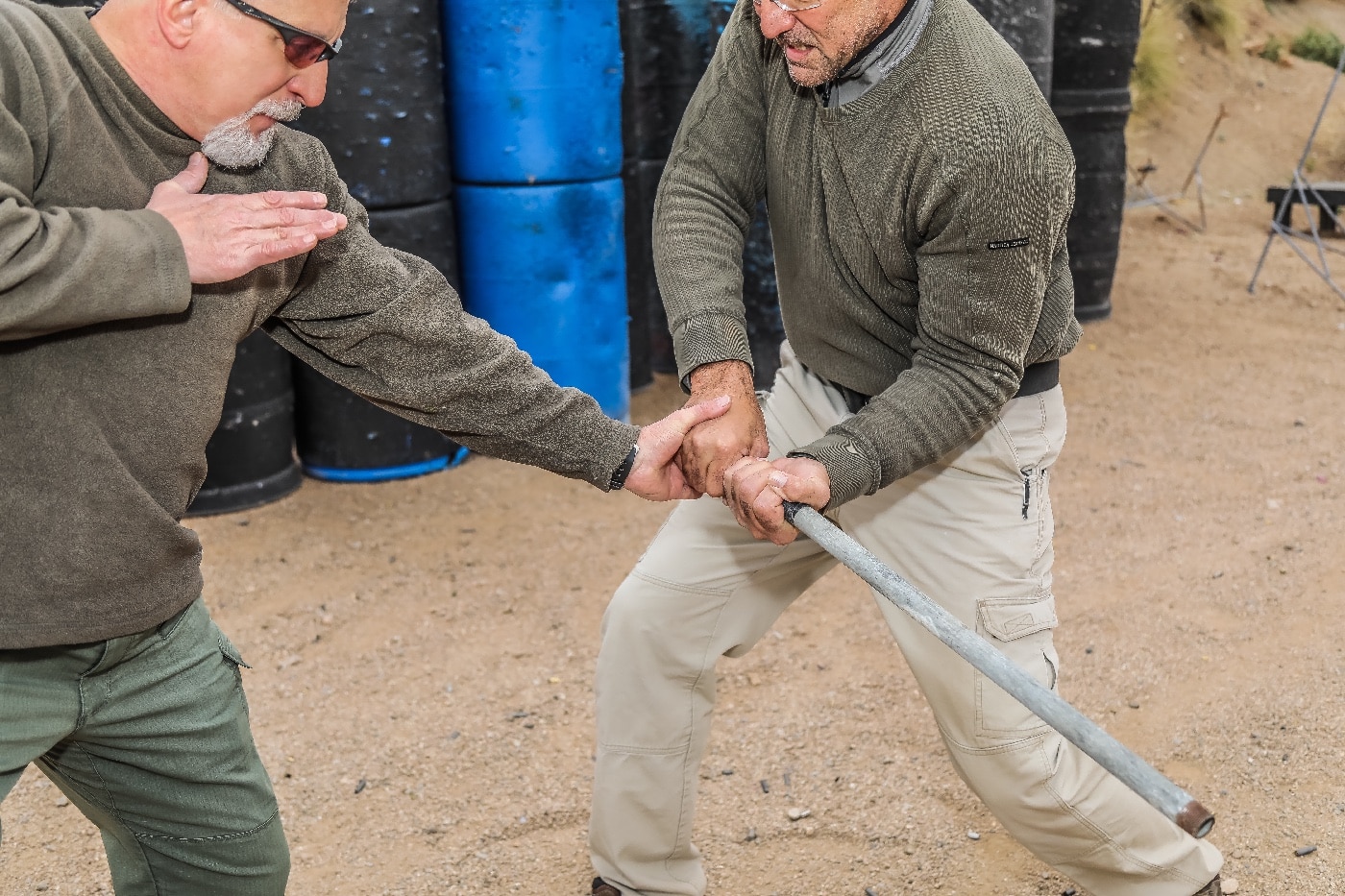 In this image, one person swings a blunt impact weapon, such as a metal pipe or club, at the knees of another individual, who is quickly shifting their leg away while lowering their center of gravity. The defender’s body position is alert and mobile, demonstrating the need for immediate reaction to protect vulnerable joints from disabling strikes. The attacker is focused on landing a blow that could end the fight by taking out the legs, highlighting how low-line attacks are a serious threat in street fights. The setting appears to be an outdoor area like a parking lot or alley, adding a realistic backdrop to the confrontation. This photo emphasizes the importance of defensive movement and lower body protection during violent encounters. Clothing and posture of both individuals show the unpredictability of real-world fights. The scene makes it clear that attacks can come from any angle and at any height, not just to the upper body.