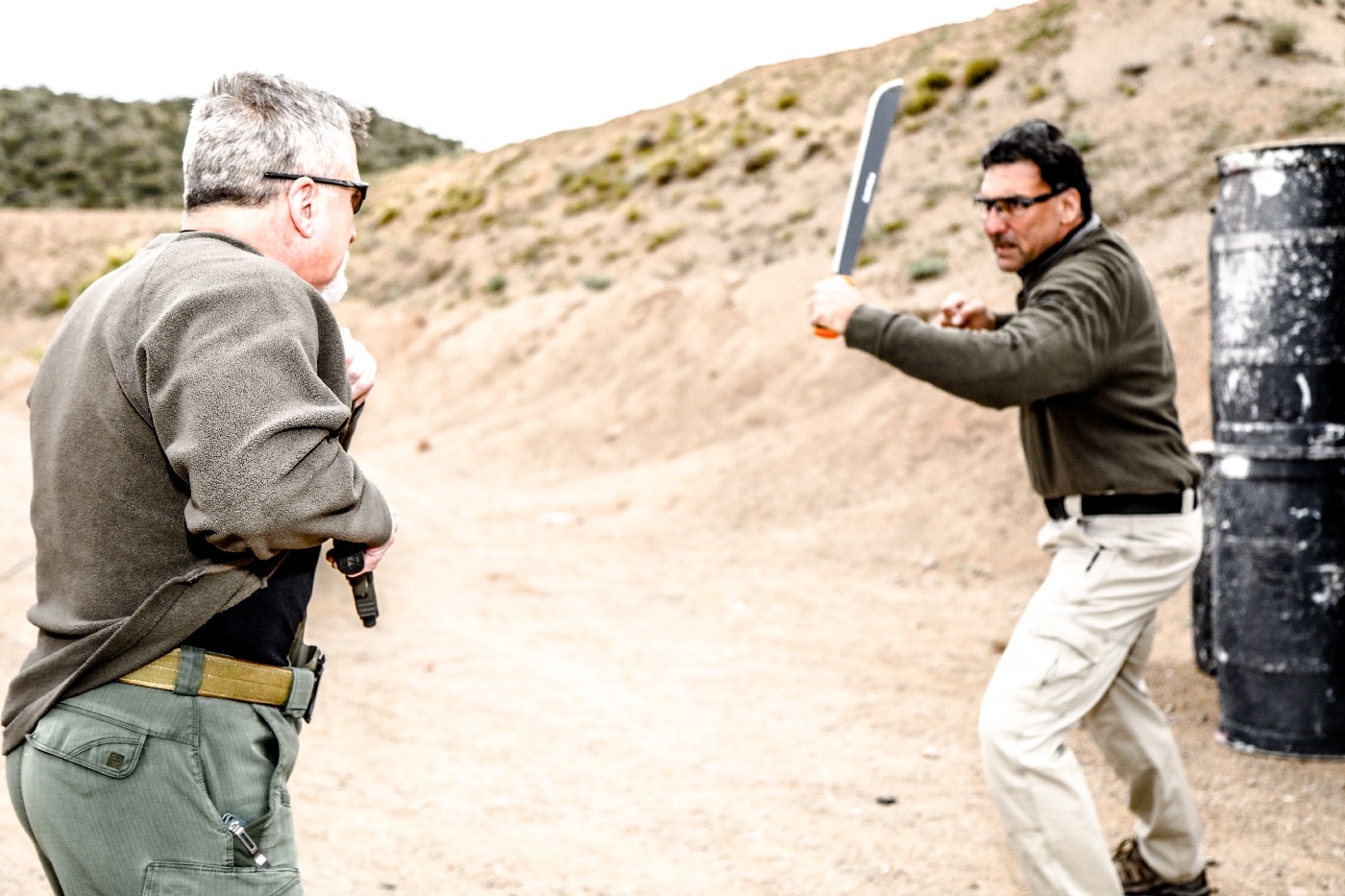The photo captures a tense moment where one individual, faced with an attacker wielding a raised machete, rapidly draws a concealed handgun to defend themselves. The defender’s movement is quick and purposeful, emphasizing the urgency of accessing their firearm under life-threatening circumstances. The assailant’s aggressive posture, machete held overhead, signals imminent danger and the potential for severe injury. The background appears to be an urban or residential street, adding to the realism and relatability of the scenario. This image supports the main keyword “winning a street fight” and integrates related terms like self-defense, firearm deployment, and weapon confrontation. The photo visually demonstrates the concept of equalization and the necessity for hard skills in lethal force encounters. The description provides clarity for visually impaired readers while focusing on the high-risk nature of armed street fights. It’s also optimized for those searching about defensive handgun use in violent altercations.