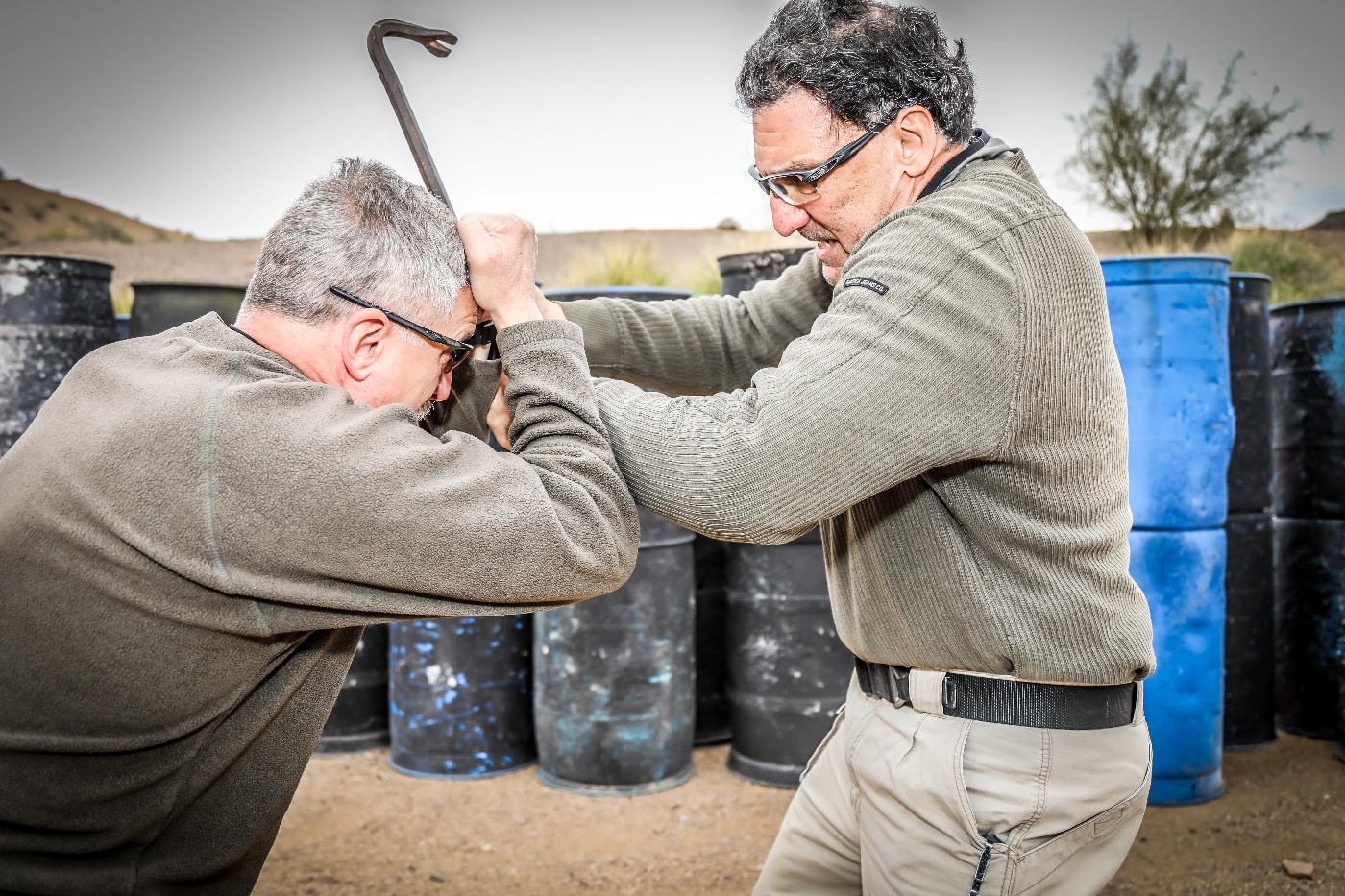The photo shows a tense moment as one person aggressively swings a crowbar at another, who raises their arms in a defensive posture to protect their head. The attacker’s body language is hostile, clearly intent on causing serious harm, while the defender is focused on survival and blocking the blow. This scene highlights the extreme danger posed when weapons are involved in street fights, reinforcing the article’s main keyword “winning a street fight” and related terms like self-defense, armed aggression, and improvised weapon. The setting is urban, possibly an alley or deserted sidewalk, with no intervention from bystanders or authorities. This description emphasizes the urgent need for movement, hard skills, and equalization when facing a lethal threat. The image serves as a stark warning about the escalation of violence in real-world altercations. It’s designed for visually impaired users seeking content on street fight defense strategies.