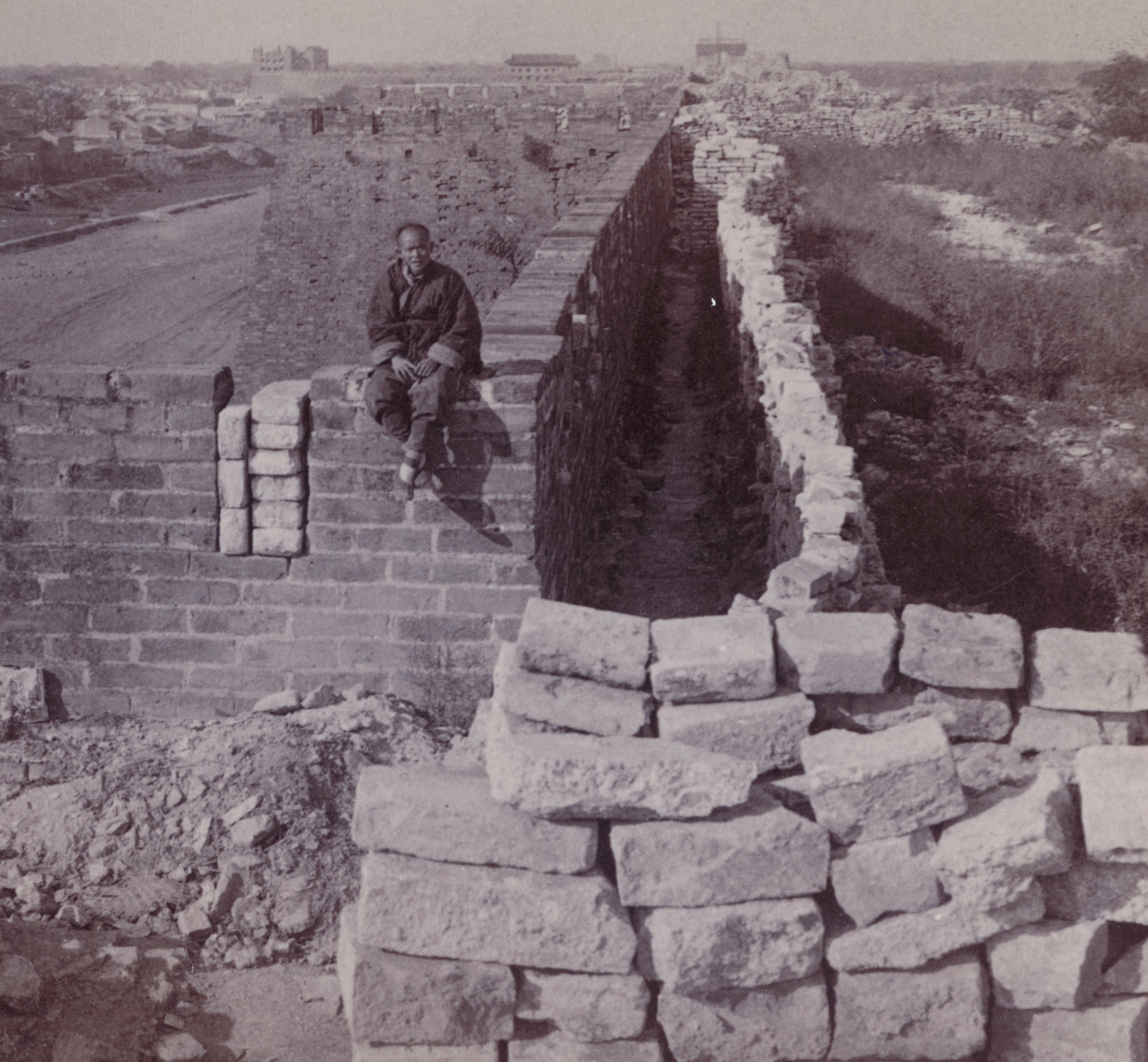 The photograph shows a broad view of the Tartar Wall as it passes near the grounds of the U.S. Legation in Peking (Beijing), China. The wall is built of massive stone and brick, towering above surrounding buildings, with crenellations and possibly a walkway along the top. In the foreground, the legation’s compound or neighboring structures are visible — low walls, courtyards, and possibly sandbagged defensive positions set up by Marines and diplomats during the Boxer Rebellion. The area is quiet but bears evidence of recent conflict, such as patched bullet holes or small guard posts. The proximity of the Tartar Wall emphasizes its strategic significance for both defenders and attackers during the 55-day siege. The photo illustrates the dramatic contrast between the ancient city fortifications and the modern reality of foreign presence in 1900 Peking.