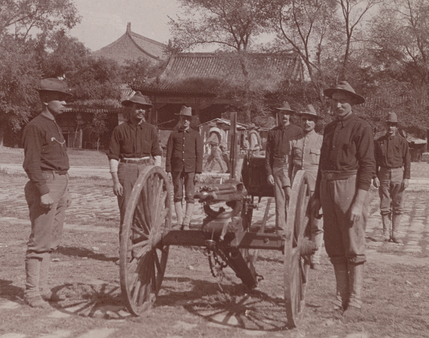 The photograph shows a group of U.S. 9th Infantry soldiers gathered around a Gatling gun during the China Relief Expedition in 1900. The soldiers are in field uniforms, some kneeling or standing with rifles slung over their shoulders, while others position or operate the multi-barreled, hand-cranked Gatling gun mounted on a wheeled carriage. The background suggests a temporary encampment, battlefield, or defensive position in China, with sandbags, makeshift barricades, or local structures nearby. The scene demonstrates how rapid-fire weapons were deployed by American troops to provide suppressive fire and protect against Boxer attacks. The soldiers’ attentive postures and the gun’s prominent placement emphasize the tactical significance of machine guns in this conflict. This image offers a clear visual of American military technology and field operations during the Boxer Rebellion.