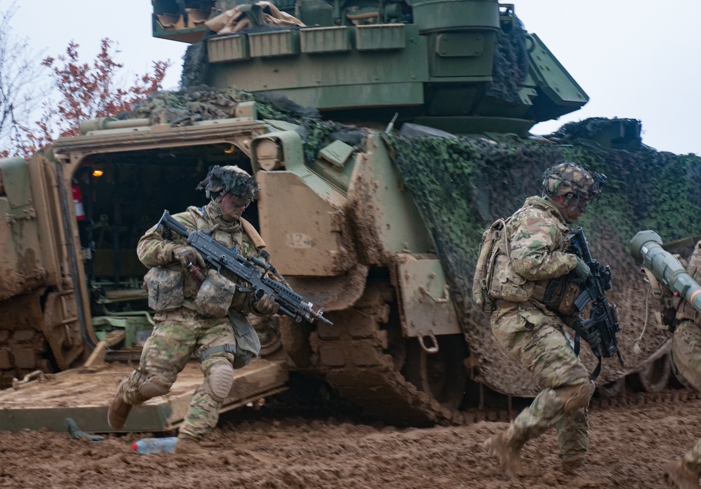 U.S. Army soldiers from the 1st Infantry Division are pictured dismounting from an M2 Bradley fighting vehicle during a live-fire qualification at Novo Selo Training Area in Mokren, Bulgaria. The photo captures several troops exiting the rear ramp of the Bradley, moving quickly onto the training ground in full combat gear, including helmets and rifles. The vehicle is positioned on open terrain, typical of a military range, with a mix of grassy and dirt surfaces in the background. The crew remains inside the Bradley, maintaining security as soldiers dismount. This training emphasizes fast, coordinated exits and immediate movement toward objectives under realistic conditions. The M2 Bradley’s rear ramp and troop compartment are visible, highlighting its function as an infantry carrier. Scenes like this are standard in multinational training events for improving combat readiness.