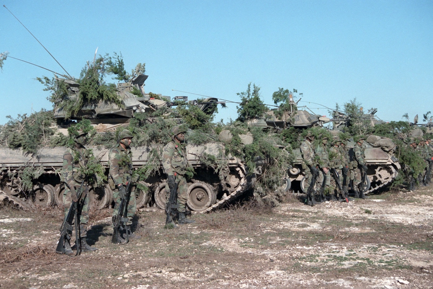 This image shows soldiers of the 2nd Armored Division standing beside their M2 Bradley infantry fighting vehicles following a company team attack exercise at the Shell Point training area. The Bradleys are covered with camouflage netting and natural foliage to blend into the surrounding environment. The soldiers, dressed in standard-issue combat uniforms and helmets, are gathered casually near the vehicles, some holding weapons, while others appear to be discussing the outcome of the exercise. The setting includes open ground, sparse vegetation, and a clear sky, typical of a large military training site. The scene illustrates post-mission regrouping, equipment checks, and team coordination after a simulated attack. Camouflage techniques are clearly visible, demonstrating practical fieldcraft. The M2 Bradley is presented as a core part of mechanized infantry operations.