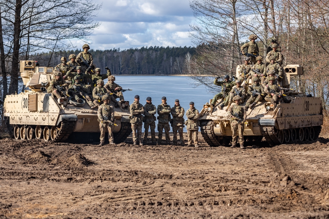 This image shows a diverse group of soldiers from the United States, United Kingdom, Croatia, and Poland posing for a group photo on top of several M2 Bradley infantry fighting vehicles at Bemowo Piskie, Poland. The soldiers, representing multiple NATO allies, are dressed in a mix of national military uniforms, some waving or smiling at the camera. The Bradley vehicles are parked in formation on a flat, open training area with grass or gravel underfoot, and a cloudy sky or wooded background typical of Poland. Flags or patches from the participating countries may be visible on uniforms or vehicles. The photo captures a spirit of camaraderie and joint readiness fostered during multinational exercises. Such moments highlight the cooperation and integration central to NATO’s presence in Eastern Europe. The scene represents both military hardware and the people behind the alliance.