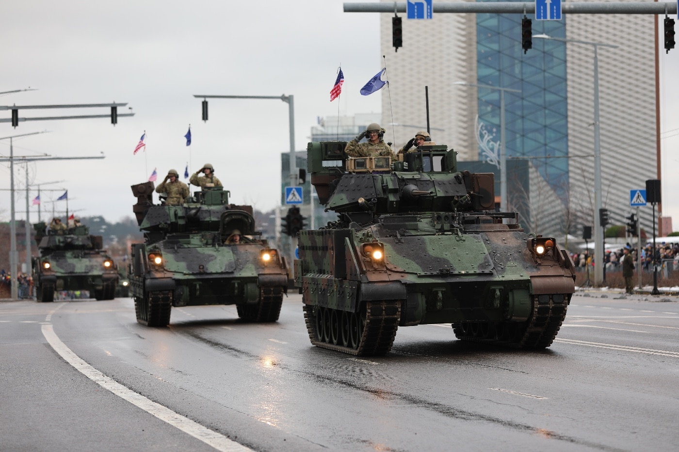 This image shows multiple U.S. Army M2 Bradley fighting vehicles driving through central Vilnius, Lithuania, as part of the 2023 Lithuanian Armed Forces Day parade. The Bradleys are painted in standard camouflage and move in a column along a main city street, passing by cheering spectators and waving Lithuanian flags. Modern city buildings and historic architecture line the route, providing a striking urban backdrop. Soldiers are visible inside or atop the vehicles, maintaining vigilance as part of the parade formation. The event is attended by civilians, families, and dignitaries, some taking photos or saluting. The parade underscores the ongoing presence and support of NATO forces in the Baltic region. The photo highlights both the impressive hardware and the symbolic importance of allied military cooperation.