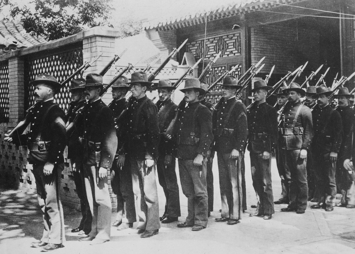 Group of U.S. Marines from the China Relief Expedition standing in formation during the Boxer Rebellion in 1900. The Marines are dressed in period-appropriate field uniforms with hats and rifles, posed in a camp or near a military installation in China. The men appear alert and ready, surrounded by makeshift equipment and supplies used during the relief mission. This image captures the American military presence that joined other international forces to defend foreign diplomats and civilians trapped in Peking. In the background, details such as tents or Chinese architecture might be visible, providing historical context. The photo illustrates the multinational effort to suppress the Boxer uprising and protect western interests in China. The Marines’ uniforms and gear highlight early twentieth-century U.S. military appearance during overseas campaigns.