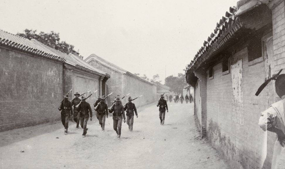 The photograph shows a group of U.S. Marines in uniform walking along a street or alleyway close to the International Legations in Peking (Beijing), China, shortly after the Boxer Rebellion. The Marines carry rifles at the ready and move in pairs or small squads, scanning the area for threats as they patrol the perimeter of the foreign diplomatic compounds. The background features a mix of Western-style legation buildings and traditional Chinese architecture, with signs of recent conflict such as damaged walls, sandbags, or barricades. Local Chinese civilians may be seen in the distance, going about daily life under the watchful eyes of the military. The image reflects the continuing security challenges and international presence in Peking following the 1900 siege. It documents the role of U.S. Marines in maintaining order and protecting foreign nationals in a city still marked by unrest.