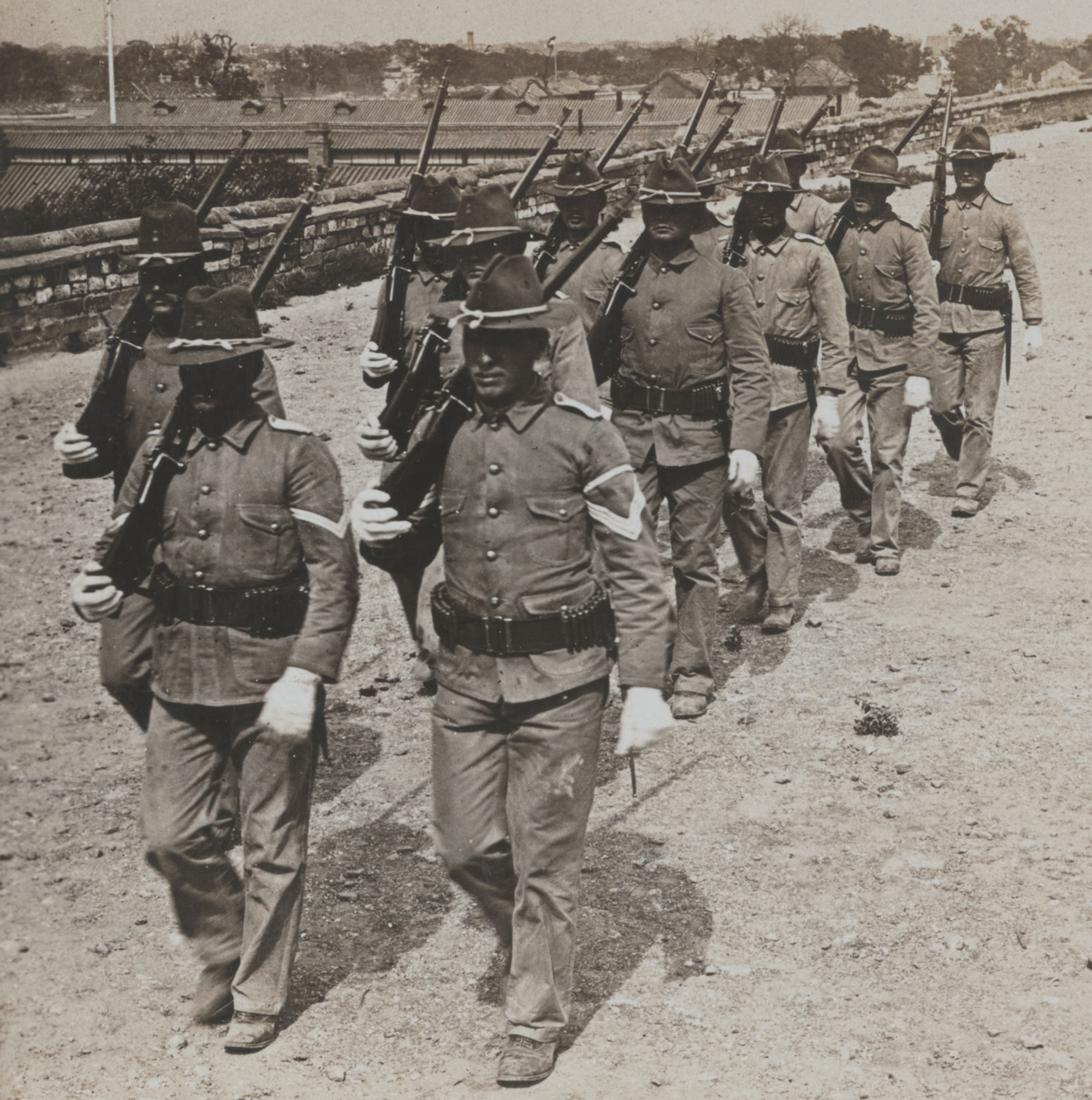 The photograph shows a column of U.S. Infantry soldiers marching in full gear along the broad, flat top of the Tartar City Wall in Peking (Beijing), China, during the Boxer Rebellion. The soldiers are spread out in a loose line, rifles slung over their shoulders or held at their sides, with some officers leading the group. The immense size of the city wall is immediately apparent: the wall stretches far into the distance, wide enough for several men to march side by side, and its height is clear from the steep drop visible on one or both sides. The background includes city rooftops, temples, or the sprawling urban landscape of Peking below, giving a sense of how high the wall rises above the surrounding area. The image conveys the strategic importance of the Tartar Wall for both defenders and attackers. The photo provides historical context for the kind of terrain faced by U.S. and allied troops in 1900 China, illustrating the monumental scale of ancient Chinese fortifications.