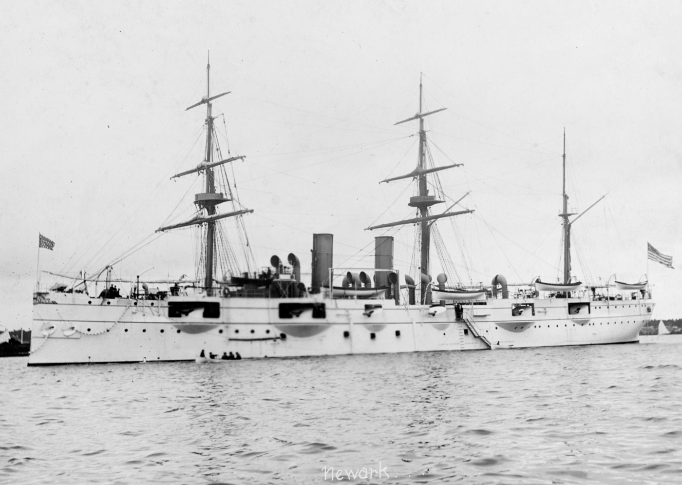 The USS Newark (C-1), a protected cruiser of the U.S. Navy, is shown anchored in calm waters, likely photographed shortly before or during her deployment to China for the Boxer Rebellion. The ship features a steel hull, prominent smokestacks, and multiple deck guns typical of late 19th-century American cruisers. Crew members can be seen on deck, and flags or signal pennants may be flying from the masts. The cruiser’s long, narrow profile and protective armored deck are evident, reflecting design priorities of the era. This image captures the Newark as she prepared to join the international naval force assembled to protect foreign interests during the Boxer uprising. The ship played a key role in landing Marines and projecting American military power in East Asia. The photo offers a detailed look at early U.S. naval technology and readiness for overseas operations.