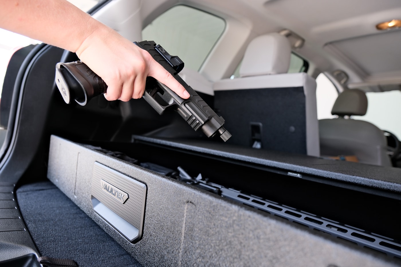 A person is shown retrieving a Springfield Armory XD-M Elite pistol from the open drawer of a Vaultek RS Slider Safe mounted inside the cargo area of an SUV. The safe is positioned for easy access, securely fastened to the vehicle’s interior. The RS Slider’s hydraulic drawer extends smoothly, revealing the handgun and other gear stored inside, all illuminated by internal LED lights. This setup highlights the safe’s flexibility as both a home and vehicle storage solution for firearms. The user’s hand reaches in to grip the XD-M Elite, emphasizing the safe’s role in providing quick, secure access even in a mobile environment. The SUV’s interior surrounds the scene, showing how the safe fits within a typical trunk or cargo space. This ALT text targets readers interested in vehicle gun safes, quick-access firearm storage, and secure transportation for handguns and rifles.