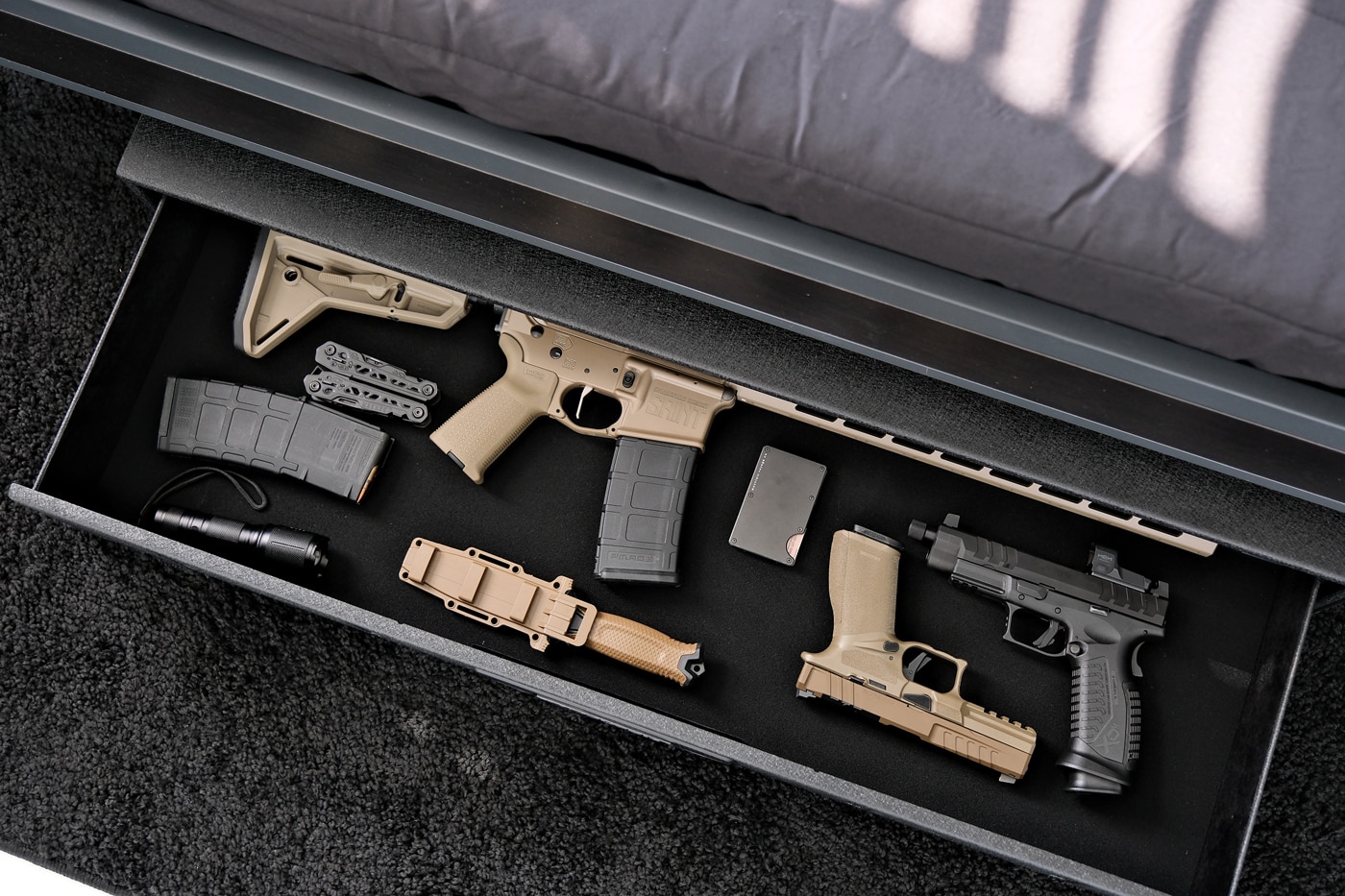 In this image, a Vaultek RS Slider Safe sits horizontally under a standard bed, with its main drawer slid open toward the camera. Inside the drawer, several home defense firearms — such as a compact AR and other accessories — are neatly stored and lit by built-in LEDs. The steel safe appears compact and is fully concealed beneath the bed aside from the drawer opening. The integrated touchpad and fingerprint sensor on the face of the safe are visible, highlighting the quick-access feature. This under-bed storage offers a secure but fast way to access firearms in a home defense scenario. The setup is suitable for anyone considering a slim rifle safe for tight spaces. The image reinforces the safe’s blend of security and convenience in personal gun storage.
