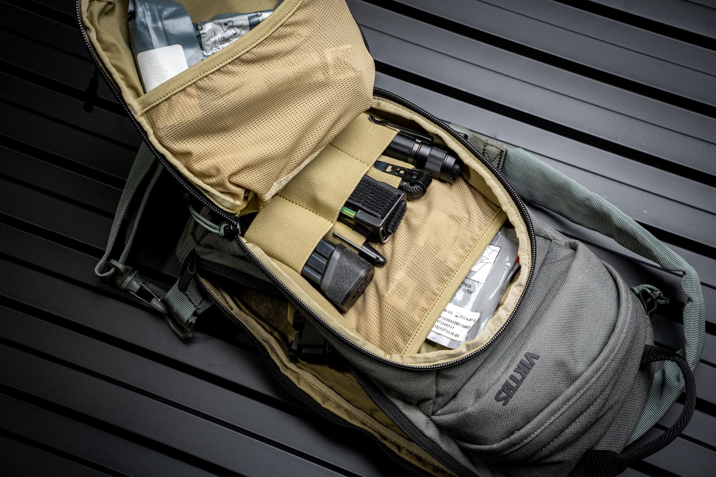 Photo of the Viktos Upscale 3 Slingbag with its main compartment fully open, displaying several spare pistol magazines, compact first aid gear such as a trauma dressing and tourniquet, and a tactical flashlight. Each item is placed in its own pocket or section, showing the bag’s ability to keep gear separated and organized. The magazines are secured in elastic holders, the first aid supplies are packed into a mesh pocket, and the flashlight is easily accessible in an adjacent slot. The tough nylon material, strong zippers, and reinforced seams of the bag are visible around the edges. This layout demonstrates practical daily carry, with all emergency and backup items ready for use. The background is clean, keeping the viewer’s focus on the contents and the compartment’s design.