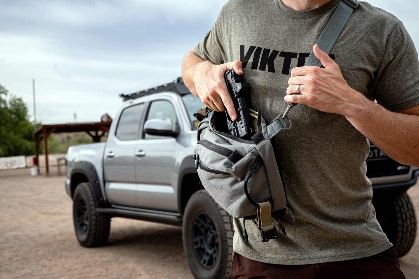 Close-up photograph showing a person’s hands as they draw a semi-automatic pistol from the concealed compartment of a grey Viktos Upscale 3 Slingbag. The bag is slung across the front of the torso and partially unzipped, allowing easy access to the firearm. The image is meant to illustrate the off-body carry process in a real-world context. The focus is on the mechanics of drawing the pistol, including grip and compartment layout. The Viktos Upscale 3’s design features — like the padded shoulder strap, tactical zippers, and Velcro-lined holster attachment — are partially visible. This is the first image in the Viktos Upscale 3 Slingbag review article and is intended to provide an overview of how the bag facilitates practical self-defense carry. The background is neutral and unobtrusive, keeping attention on the bag and the author’s hands. The photo is useful for visually impaired users wanting to understand the hands-on function of this sling bag for concealed carry.