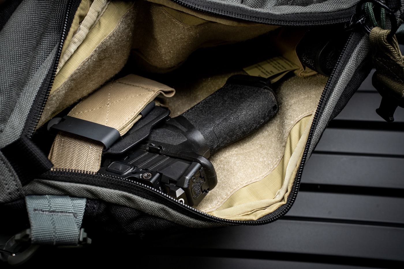 Photograph of the Viktos Upscale 3 Slingbag with its concealed carry compartment fully unzipped, displaying a pistol held in a dedicated holster secured to the Velcro-lined interior. The firearm is positioned grip-up for right-handed access, with the holster firmly attached to prevent shifting during movement. The compartment is designed to keep the pistol separate from other bag contents, reducing the risk of accidental contact or interference. The sturdy zippers and reinforced compartment walls are clearly visible, emphasizing the bag’s build quality and safety features. The bag is resting on a neutral background, with all attention focused on the concealed carry setup. No hands or accessories are in the frame, making it easy to see the storage arrangement. This image is used to give readers a clear visual of how the Viktos Upscale 3 supports concealed carry for daily use.