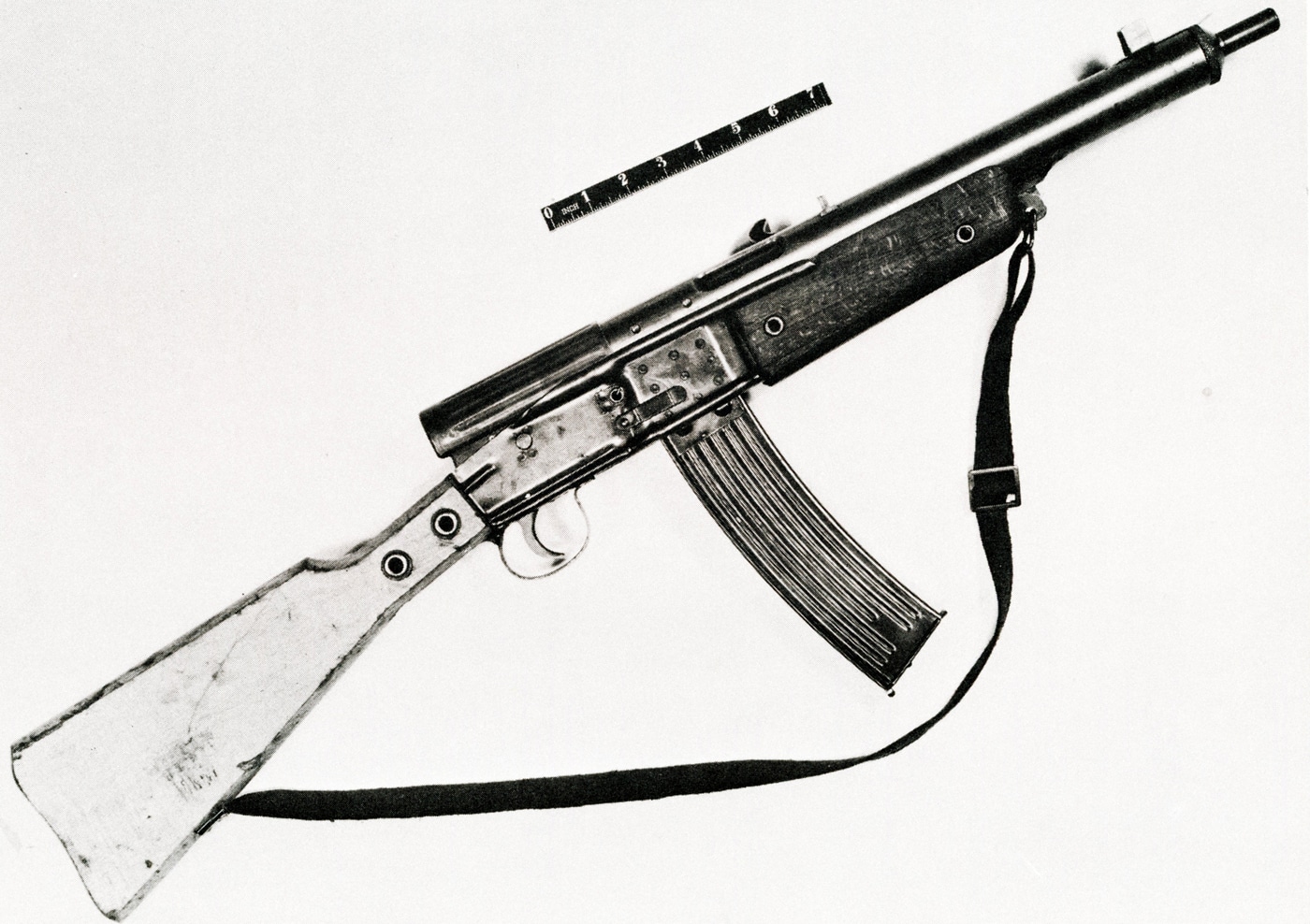 The image shows a Gustloff Volkssturmgewehr rifle displayed in a glass case or on a mount within the U.S. Army Ordnance Museum collection. The rifle is positioned for public viewing, with its simple stamped metal receiver, fixed wooden stock, and prominent curved magazine clearly visible. Lighting in the display area highlights the utilitarian construction and crude finishing typical of late-war emergency firearms. Informational placards or exhibit labels may be present nearby, offering historical context about the weapon’s origin and use. The surrounding museum environment includes other military artifacts and firearms, but the focus remains on the Volkssturmgewehr as a significant example of German World War II arms. This preserved piece allows visitors to study the features and materials used during Germany’s desperate push for mass production. The display helps illustrate the technological and logistical realities faced by the German military in the closing months of the conflict.