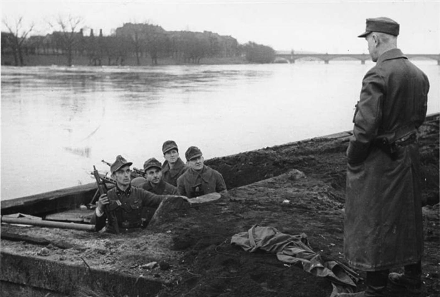 The image shows a group of Volkssturm soldiers in uniform, stationed along the banks of the Oder river during the final stages of World War II. Each soldier is armed with a Gustloff Volkssturmgewehr rifle, a distinctive weapon known for its crude construction and late-war production. The soldiers are using natural cover such as shrubs and earthworks, reflecting the defensive preparations common in 1945 as Soviet forces closed in on Berlin. The Gustloff Volkssturmgewehrs in their hands are easily identifiable by their box magazines and simple, utilitarian design. The overall mood is tense and somber, illustrating the desperation of Germany’s last-ditch efforts to resist invasion. The terrain and river setting emphasize the historical location and significance of the Oder front in the war’s closing weeks. This photograph documents both the appearance of Volkssturm troops and their reliance on emergency-issue firearms like the Volkssturmgewehr. The image is a key visual record of the conflict’s final defensive battles.