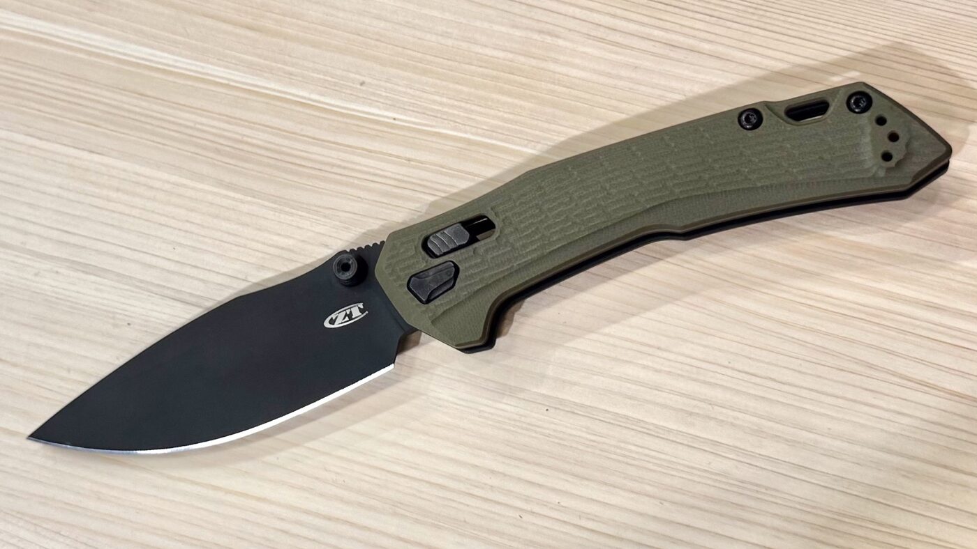 This image shows the Zero Tolerance 0203 folding knife on display at the 2025 Blade Show with its blade open. The knife features a CPM M4 steel blade, coated in black PVD, and has a pronounced leaf-shaped drop point profile. The handle is constructed from textured olive green G10 and appears thick and ergonomic for a secure grip. The open blade and handle are both sharply defined, and the Duralock mechanism is visible near the pivot. The knife rests on a branded Zero Tolerance display mat or table, with other ZT knives and promotional materials possibly visible in the background. The scene is brightly lit, emphasizing the knife’s robust construction and modern tactical design.