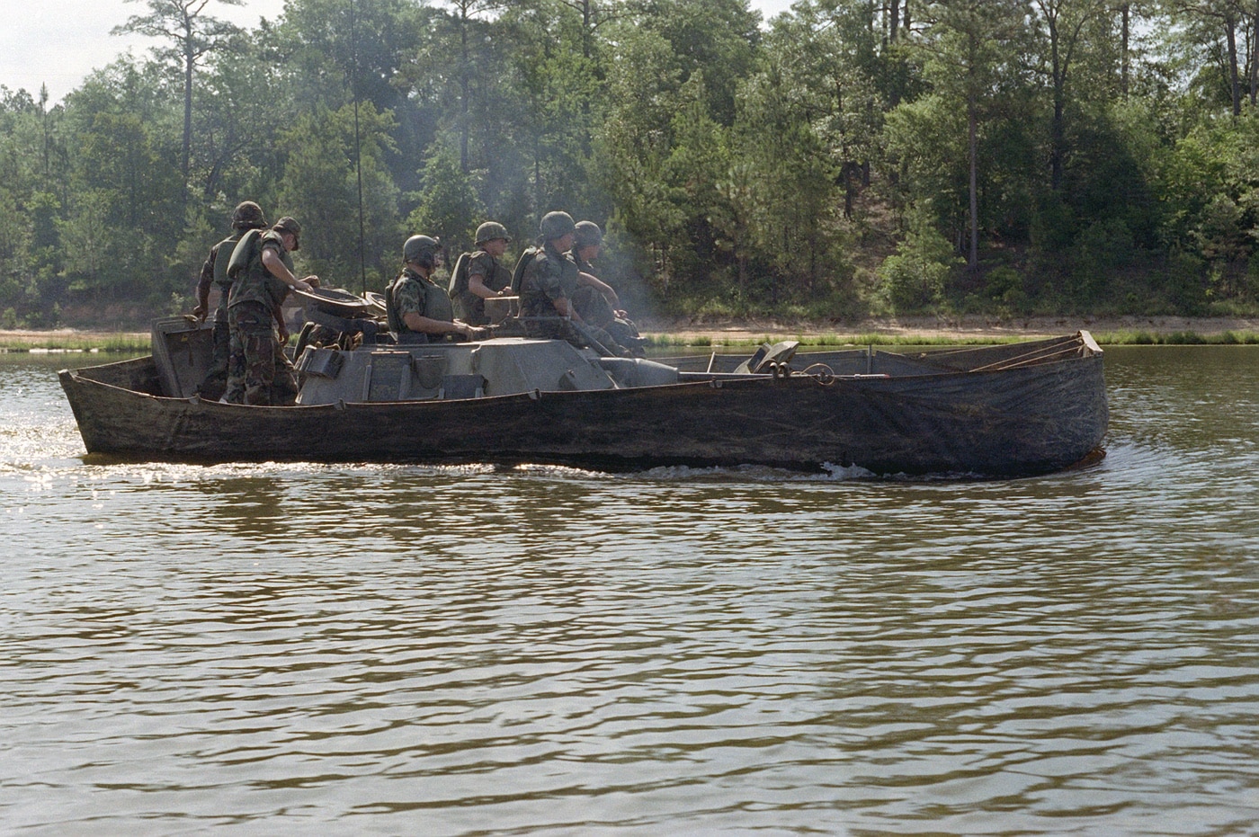 The image shows a group of infantrymen training with an M2 Bradley infantry fighting vehicle in the water, demonstrating the vehicle’s amphibious capabilities. The Bradley is partially submerged as it crosses a pond or small lake, with water reaching up to the lower hull. Crew members are visible on or inside the vehicle, monitoring its movement and performance during the exercise. The M2 Bradley’s tracks, which act as the primary means of propulsion in water, are clearly visible beneath the surface. The setting includes calm water with grassy or muddy banks and trees in the background, typical of a military training area. This exercise allows soldiers to learn proper procedures for launching, navigating, and exiting the water. The scene illustrates a less commonly used but important capability of the Bradley for river or obstacle crossings.