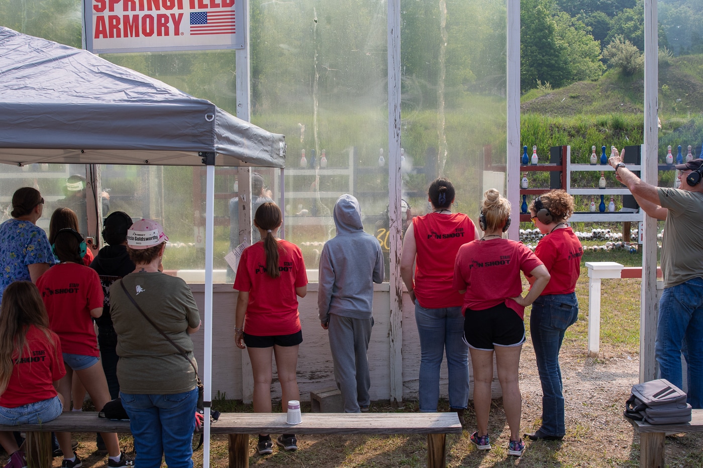 The image shows several timekeepers standing by the firing line during an active round at the Springfield Armory Shootoff. The timekeepers, identifiable by their stopwatches or timing devices, are positioned just behind or to the side of the shooters, carefully monitoring each run. In the foreground or mid-ground, competitors can be seen in shooting stances, aiming and firing at the bowling pin targets downrange. The timekeepers’ focused expressions and attentive body language highlight their responsibility for recording accurate times. The background includes the outdoor range environment, with shooting tables, pin arrays, and possibly audience members in the distance. Safety gear such as hearing protection and eye protection is visible on both shooters and officials. This photo emphasizes the critical support role that timekeepers play in maintaining the integrity of the match.