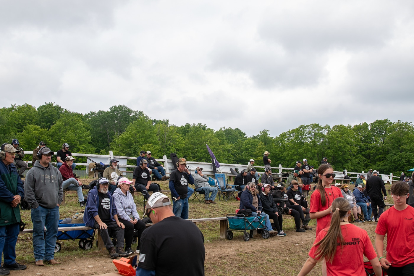 The photo shows the audience beginning to fill the bleachers set up on a grassy hillside behind the firing line at the Springfield Armory Shootoff. People of different ages are taking their seats, some holding refreshments or programs, while others chat and look toward the range. The setting is outdoors, with natural light and green surroundings typical of Central Lake, Michigan. A few spectators are already seated, while others are still arriving or climbing the steps. The bleachers provide an elevated view of the competition area, with shooting tables and targets visible in the distance. The crowd includes both families and individual fans, creating a lively and anticipatory atmosphere. This image highlights the rare spectator-friendly design of the Pin Shoot and the community interest in the event.