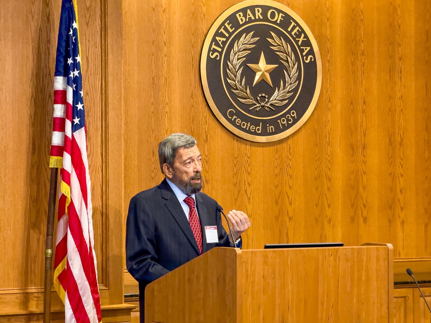 The photo shows Massad Ayoob standing at a podium in a conference room, speaking to a group of attorneys during a Continuing Legal Education seminar sponsored by the State Bar of Texas. He gestures while addressing the audience, who are seated and taking notes or listening attentively. The setting is professional, with presentation materials and legal documents visible on tables or screens. This scene highlights the importance of education for lawyers on self-defense trial strategy and how firearms training factors into legal defense. The image is relevant to topics about legal education, self-defense law, and the ways attorneys prepare for defending self-defense shootings in court.
