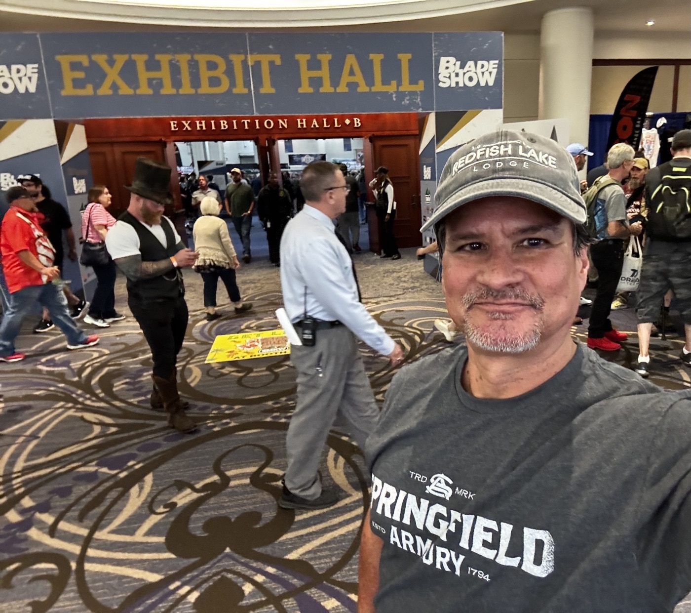 This photo shows the author standing at the entrance of the 2025 Blade Show in Atlanta, Georgia. The entrance to the Cobb Galleria is visible, with banners and signs indicating the event and its dates. There are groups of attendees and visitors gathered near the doors, many holding bags or passes, ready to enter the venue. The setting is busy, capturing the excitement and anticipation of opening day at the world’s largest knife show. The author is clearly visible, possibly holding a badge or ticket, and is dressed casually for the event. Large glass windows and modern architecture are part of the background, and there may be promotional posters or Blade Show branding displayed nearby. This image helps set the scene for an article focused on the 2025 Blade Show and the new knives featured at the event.