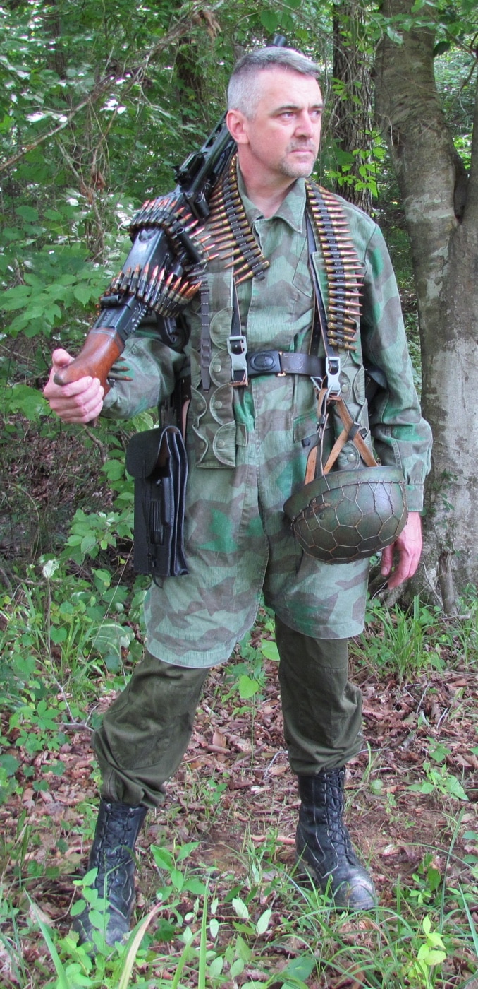 A photo of the article’s author walking with an MG 42 machine gun, either slung over the shoulder or held in both hands. The gun’s receiver, barrel, and bipod are easy to see, and the author’s modern dress makes it clear this is a demonstration, not a historical image. The MG 42 appears heavy and awkward to carry, showing why it was usually a team weapon. The scene is straightforward, focusing on the physical aspect of transporting the MG 42 machine gun.