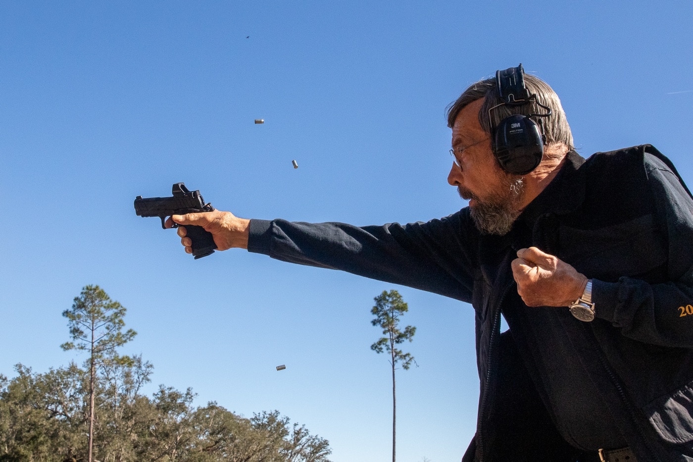 The photo shows the author at a shooting range demonstrating the trigger slap technique with a single action Prodigy pistol. The author’s trigger finger is caught in motion, springing off the trigger right after firing, while the pistol is held firmly with both hands. Personal protective equipment, such as ear and eye protection, is visible. The background includes range markers and a standard target setup, but the focus stays on the author’s hand, grip, and the fast, aggressive finger movement that defines trigger slap. This image emphasizes the use of trigger slap for rapid fire, especially at close distances, as described in the article. The author’s posture and grip show control despite the lack of finesse in finger movement. The visual supports the point that trigger slap is a valid tool when speed is more important than precision.