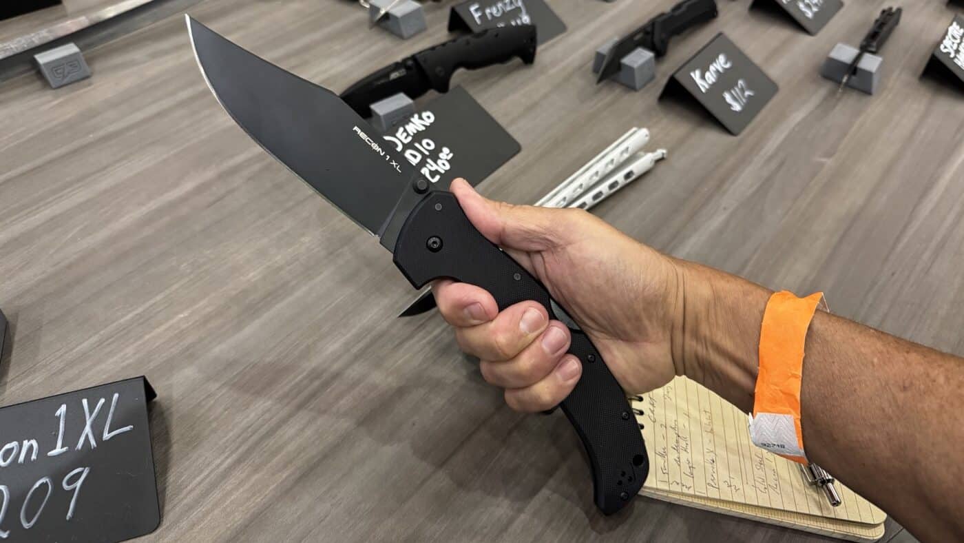 This photo shows the author holding the Cold Steel Recon 1 XL knife at the 2025 Blade Show in Atlanta. The author grips the large folder with both hands, demonstrating the impressive length of the 5.25-inch CPM XHP blade. The open knife clearly displays its clip point blade and textured G-10 handle, making the scale and construction of the Recon 1 XL obvious. The background includes the Cold Steel booth, with display tables, branded signage, and possibly other knives visible. The author appears focused on the knife, emphasizing its weight and ergonomic grip. The lighting is bright and the setting is busy, reflecting the excitement and engagement typical at Blade Show.