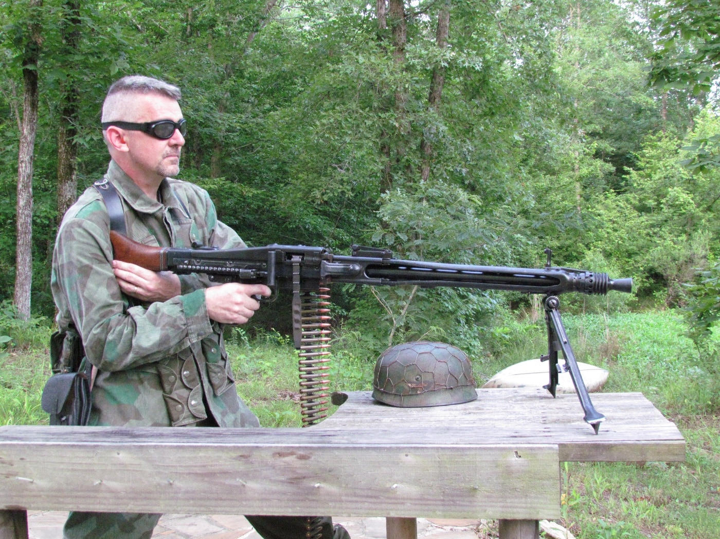 The author is shown in the act of firing the MG 42 machine gun, standing or prone behind the weapon with hands gripping the trigger and grip assembly. The MG 42 is actively cycling, with a visible blur at the ejection port or flying brass, making it clear the gun is being fired at speed. The author wears modern hearing protection and has a focused, intense expression while handling the weapon. A belt of ammunition feeds into the MG 42, and spent casings litter the ground nearby. The photo captures the real-life experience of shooting the MG 42 machine gun, known for its rapid rate of fire and strong recoil.