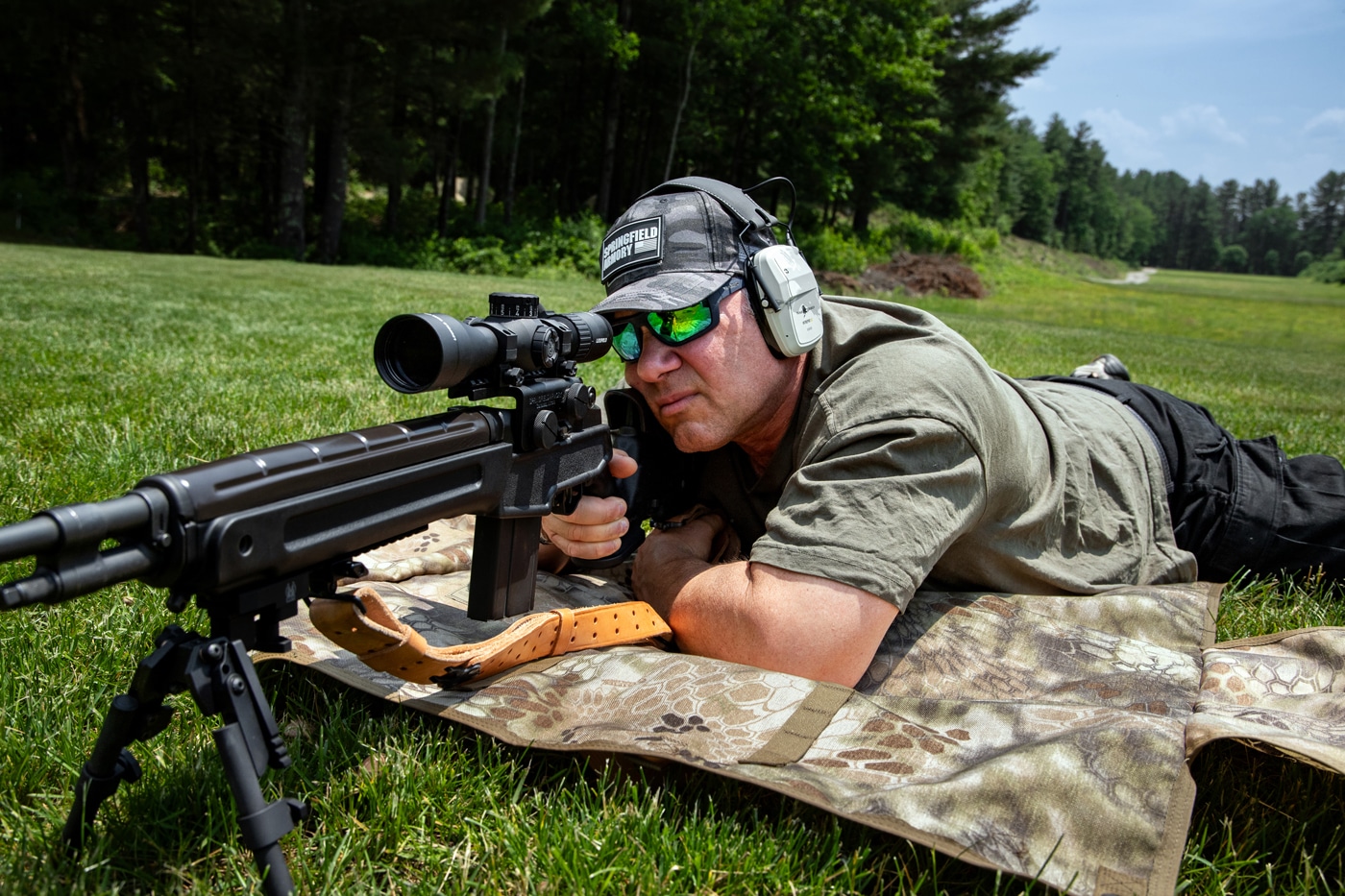 A shooter is lying prone at an outdoor shooting range with an M1A rifle, using a Leupold Mark 4HD 1.5-10x scope. The author is focused through the optic, testing how the scope performs in various lighting and distance scenarios. Spent casings and range markers are visible, indicating live-fire evaluation. This hands-on approach lets the shooter assess optical clarity, reticle visibility, and turret adjustment in a real environment. The scene highlights the Mark 4HD’s use in practical, mid- to long-range shooting situations.
