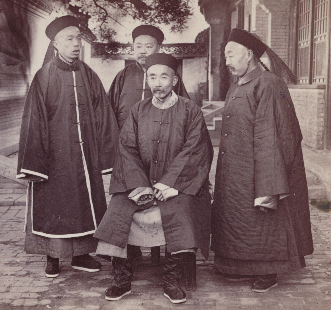 A group of senior counsellors and advisers to Empress Dowager Cixi is shown in a formal setting, possibly within the Forbidden City or another Qing palace. The men are dressed in elaborate Qing dynasty official robes, complete with rank badges, hats, and court accessories indicating their high status. Some are seated while others stand behind, posed for an official portrait or photograph, their expressions serious and composed. The setting may include richly decorated screens, carved furniture, or other elements of imperial decor. These counsellors were responsible for providing strategic and political advice to the Empress Dowager, particularly during the turbulent years of the Boxer Rebellion. Their influence was critical as the Qing court navigated internal threats from the Boxers and external pressure from foreign powers. This image illustrates the hierarchy and ceremony of the late imperial Chinese government at a moment of great upheaval.