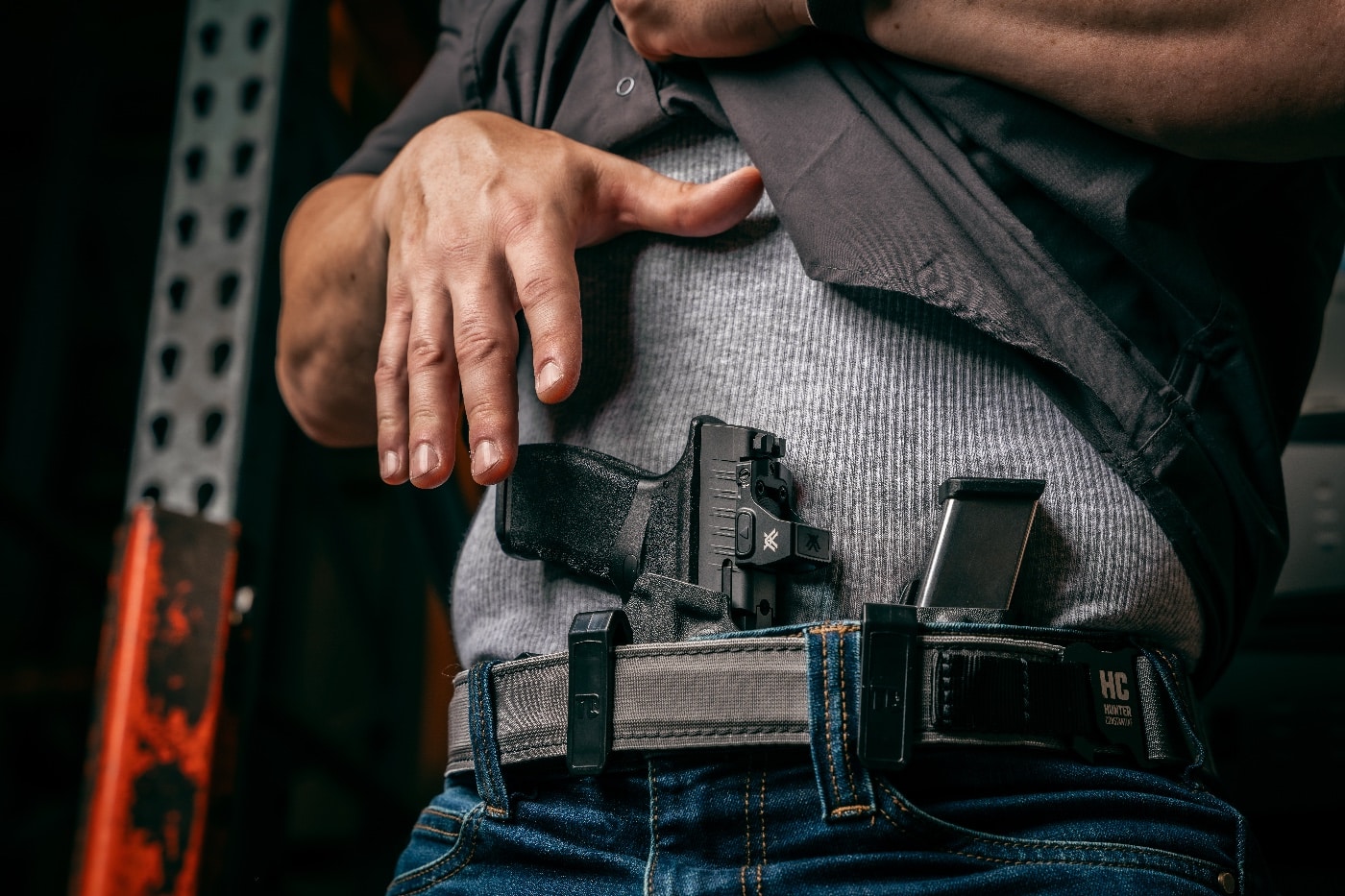 A man quickly draws a Springfield Armory Hellcat pistol from an appendix holster at the front of his waistband. His movement is focused, with one hand lifting his shirt and the other gripping the handgun securely. The man’s body language shows urgency and alertness, responding to a perceived threat. This is a common defensive draw technique used for fast access in emergencies. Recognizing dead giveaways you’re about to be attacked can lead to moments like this, where fast action is critical. The photo demonstrates both preparation and awareness in a self-defense scenario.