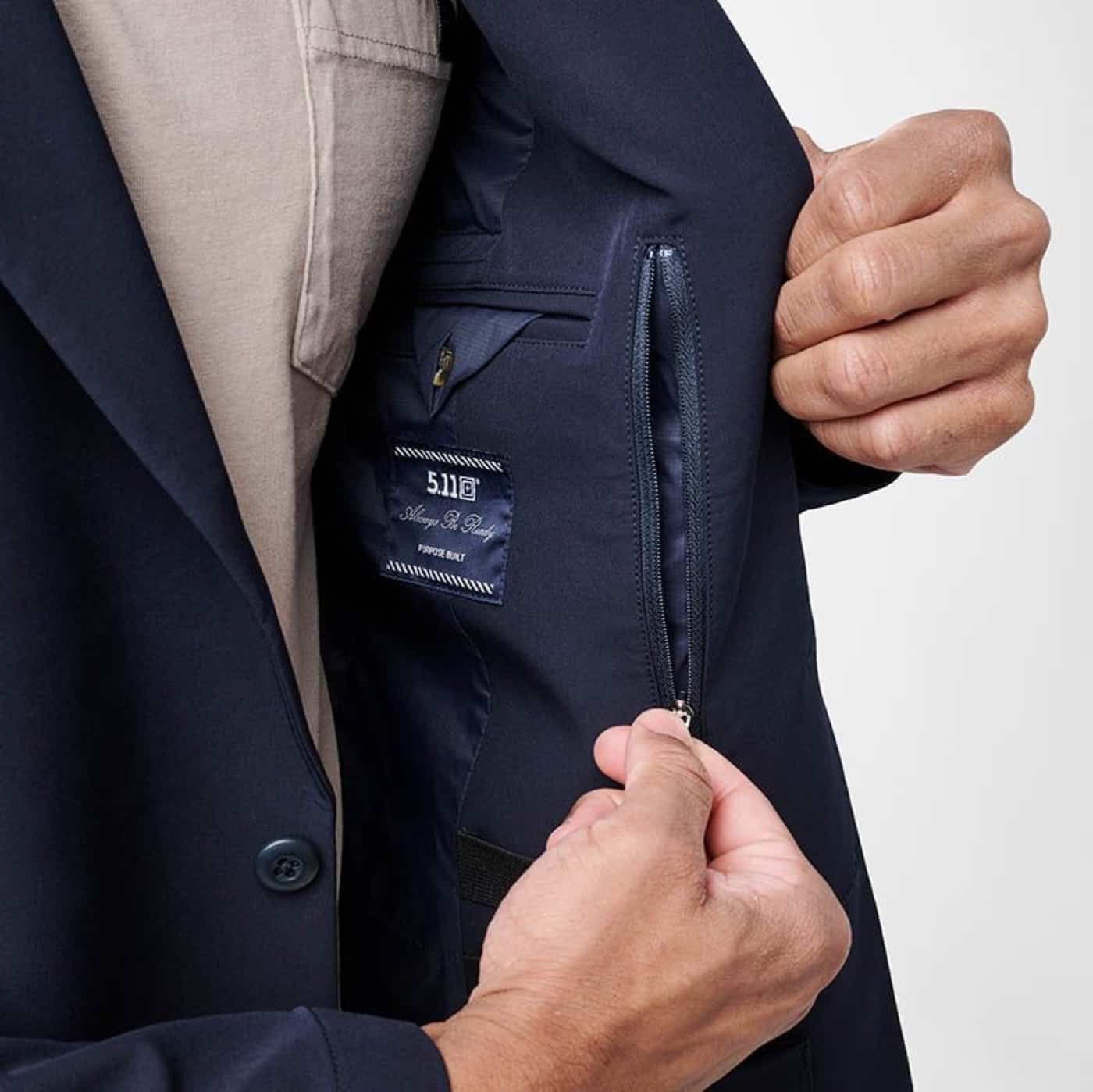 The photo shows the author pulling open one side of the 5.11 Founder’s Jacket to demonstrate the interior zippered pocket. The author’s hand is clearly holding the jacket open so the pocket is easy to see, with the zipper partially open to show its size and placement. The pocket is sewn into the lining on the inside chest area, positioned for secure but fast access to items like a phone, wallet, or small tools. Additional pockets and loops are partially visible around the zippered pocket, emphasizing the organized storage features. The jacket itself maintains a clean, professional exterior while offering tactical advantages inside. This image is used in a 5.11 Founder’s Jacket review to help readers understand the practical, user-friendly design for concealed carry or security work.