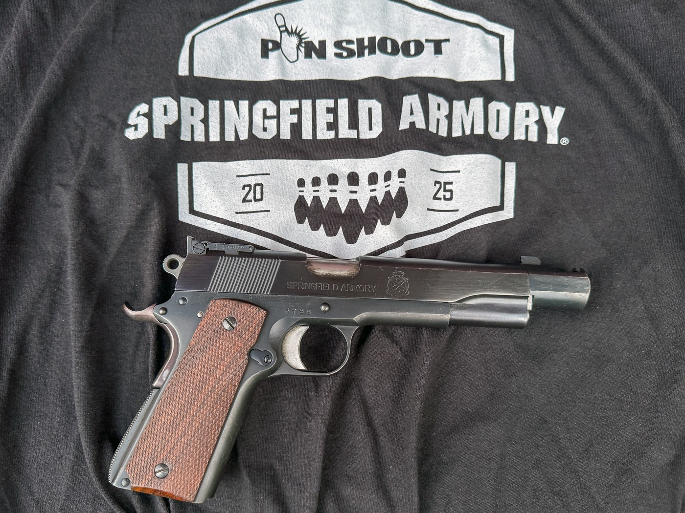 A close-up photo shows the official Springfield Armory Pin Shoot event T-shirt, laid out flat with a custom Springfield Armory 1911 pistol resting on top of it. The black T-shirt features a unique design or logo specifically created for the Shootoff, identifying it as exclusive event apparel. The custom 1911 handgun is set up for bowling pin shooting, with features such as an extended magazine, upgraded sights, and a recoil compensator clearly visible. The gun is positioned to highlight its modifications and the Springfield Armory branding. The image is taken in good lighting, emphasizing both the shirt’s design and the firearm’s details. This photo connects the signature apparel with the specialized equipment used in the match. Both items are central symbols of the 2025 Springfield Armory Shootoff experience.