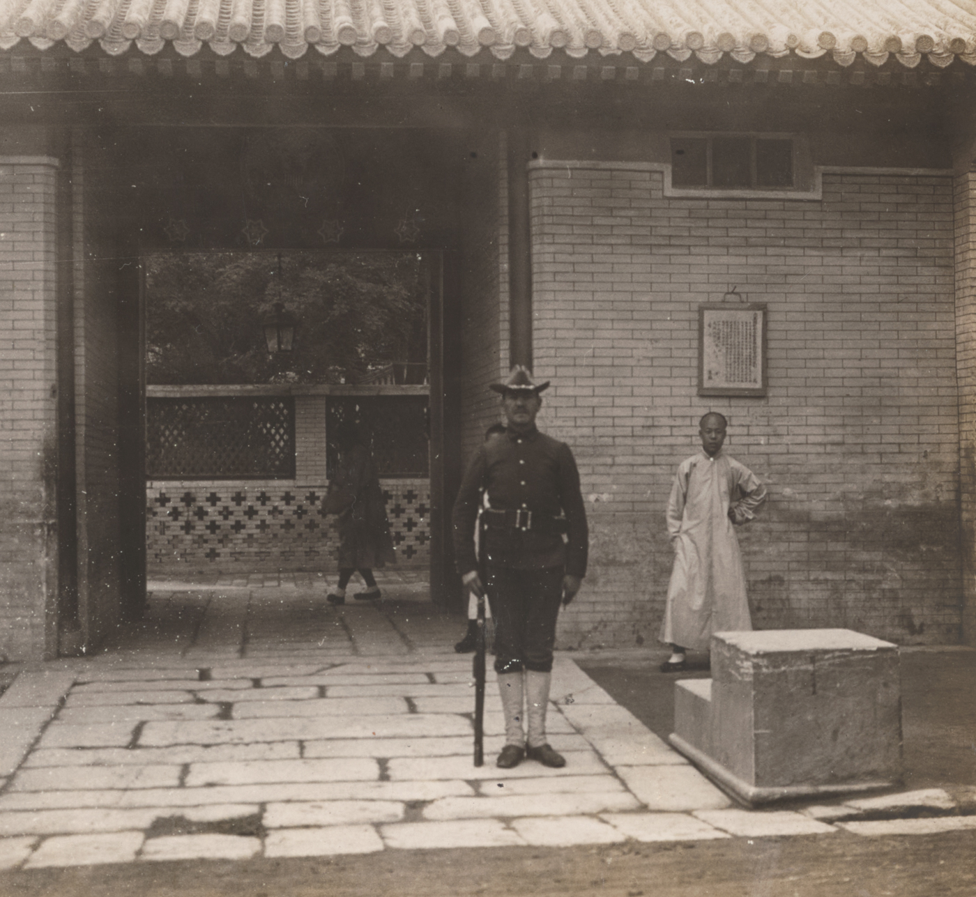 The photograph shows the entrance to the American Legation in Peking (Beijing), China, during the Boxer Rebellion. The main gate is framed by sturdy brick or stone walls, possibly reinforced with sandbags, wooden barricades, or makeshift defenses erected in response to the siege. American flags or signs indicating the legation’s presence are visible above or beside the entrance. A group of U.S. Marines or armed guards stands watch, dressed in period uniforms and equipped with rifles. The approach to the gate may be crowded with anxious civilians, Chinese Christian refugees, or foreign diplomats seeking safety inside the compound. Architectural features such as tiled roofs, carved lintels, or Chinese script on nearby buildings indicate the legation’s location in the heart of old Peking. The overall atmosphere is tense, with evidence of recent attacks or ongoing preparations for defense. The image documents a critical site in the international defense of the Legation Quarter during the 55-day siege of 1900.