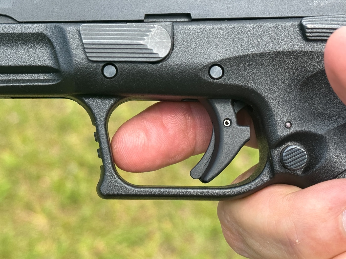 The photo shows a close-up view of a shooter’s hand holding a semi-automatic pistol, with the index finger fully extended outside the trigger guard and away from the trigger. The trigger itself is all the way forward, indicating the “at rest” position discussed in Massad Ayoob’s article on trigger control techniques. No contact is made with the trigger, making it clear that the pistol is ready to start the shooting sequence. The safety gear such as gloves, safety glasses, or the range environment might also be visible in the background. This visual highlights the most basic trigger position, critical for understanding the mechanics before learning more advanced techniques. Emphasizing safety, the finger-off-the-trigger stance is often stressed in all firearms training. This photo supports Ayoob’s points about different positions along the bell curve of trigger techniques.