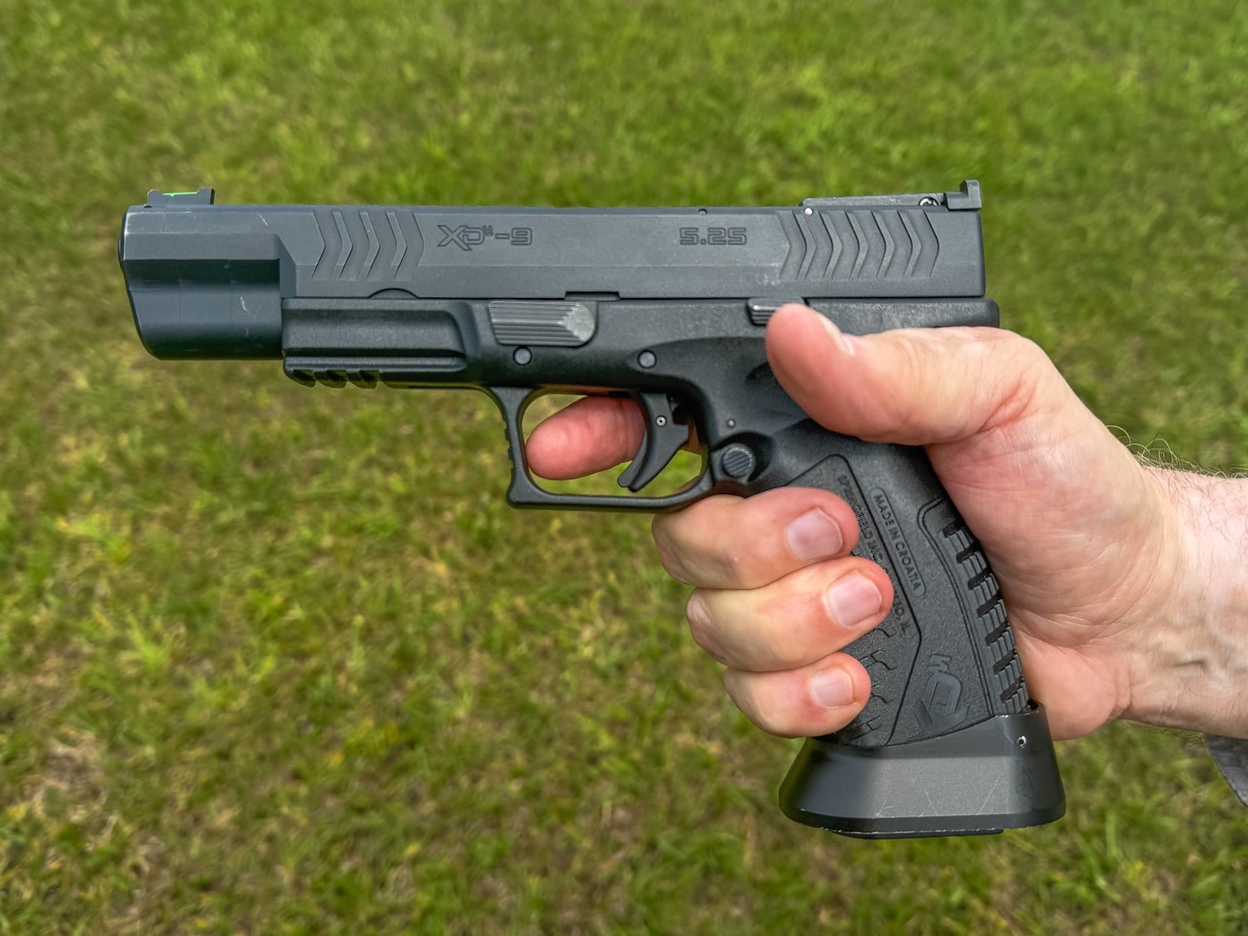 The photo shows a close-up of a shooter holding a pistol with their trigger finger straight and clearly positioned outside the trigger guard. This demonstrates proper safety protocol and trigger discipline, as the finger does not touch the trigger until the shooter is ready to fire. The rest of the hand maintains a firm grip on the firearm, and safety equipment such as eye protection is visible. The range or training area in the background reinforces the structured environment. This image directly supports the article’s emphasis on good habits for trigger control, as discussed by Massad Ayoob. The clear, visible finger-off-the-trigger stance highlights the importance of not placing the finger inside the guard until aiming and prepared to shoot. The visual is suitable for educational use, illustrating a key aspect of firearms safety and technique.
