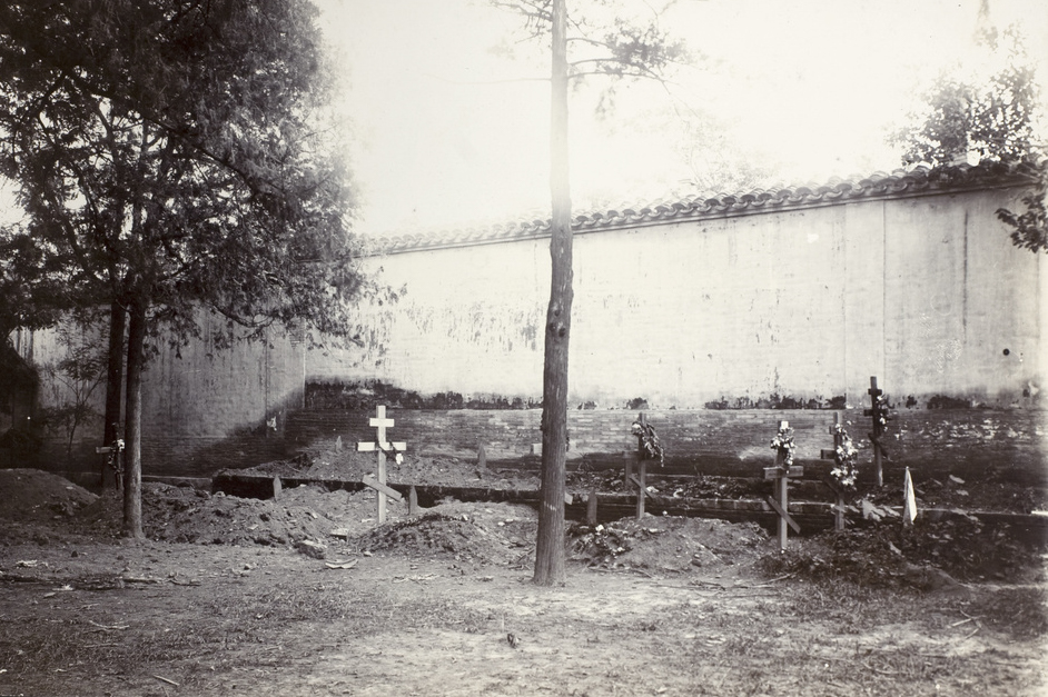 The photograph shows a small, somber cemetery within the grounds of the Russian Legation in Peking (Beijing) following the Boxer Rebellion. Several freshly dug graves are marked by simple wooden crosses or makeshift markers, each bearing hand-painted or inscribed names or units — some for U.S. Marines, others for Russian soldiers who died defending the legations during the siege. The burial plots are arranged in neat rows or small clusters, surrounded by patches of bare earth and tufts of grass. Flowers, wreaths, or flags may be placed on some graves, left by grieving comrades or diplomats. In the background, the walls and structures of the Russian Legation compound provide context for the setting. The image captures the cost of international cooperation in the face of violence and highlights the personal losses endured by both American and Russian forces during the 55-day siege.