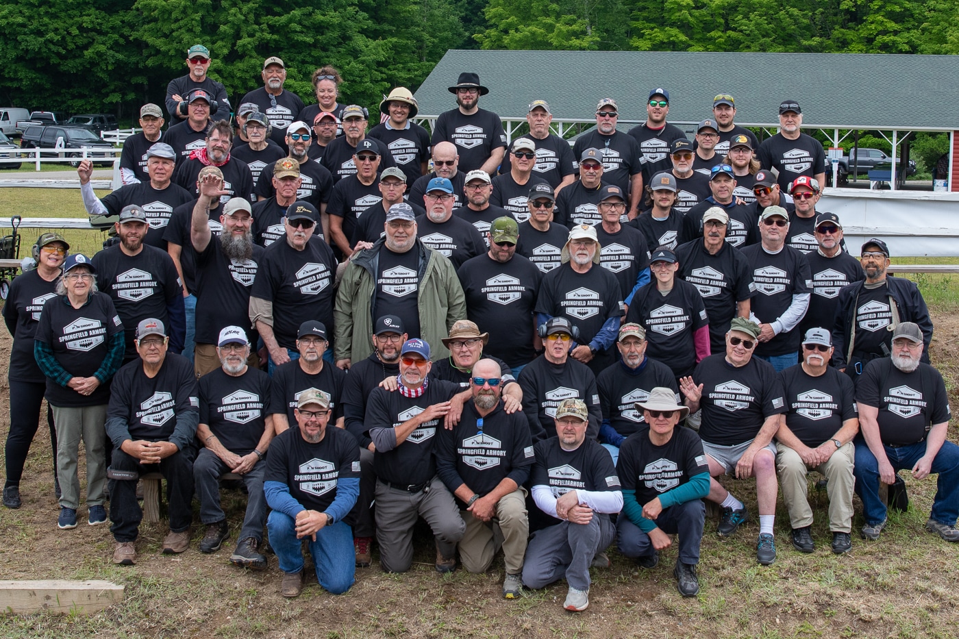 The image shows a large group of contestants assembled for a group photo at the Springfield Armory Shootoff. Participants of different ages, genders, and backgrounds are standing or kneeling in several rows, most wearing the official event T-shirt or other casual competition attire. The group is outdoors, likely in front of the shooting range or under a clear sky, with trees and range structures visible in the background. Some contestants are holding their pistols safely pointed down, while others have their arms around fellow shooters. Everyone is smiling or looking relaxed, reflecting the friendly atmosphere of the event. This group photo documents the diverse field of shooters who competed in the 2025 match. The image highlights the inclusive, community-focused nature of the Pin Shoot and Springfield Armory Shootoff.